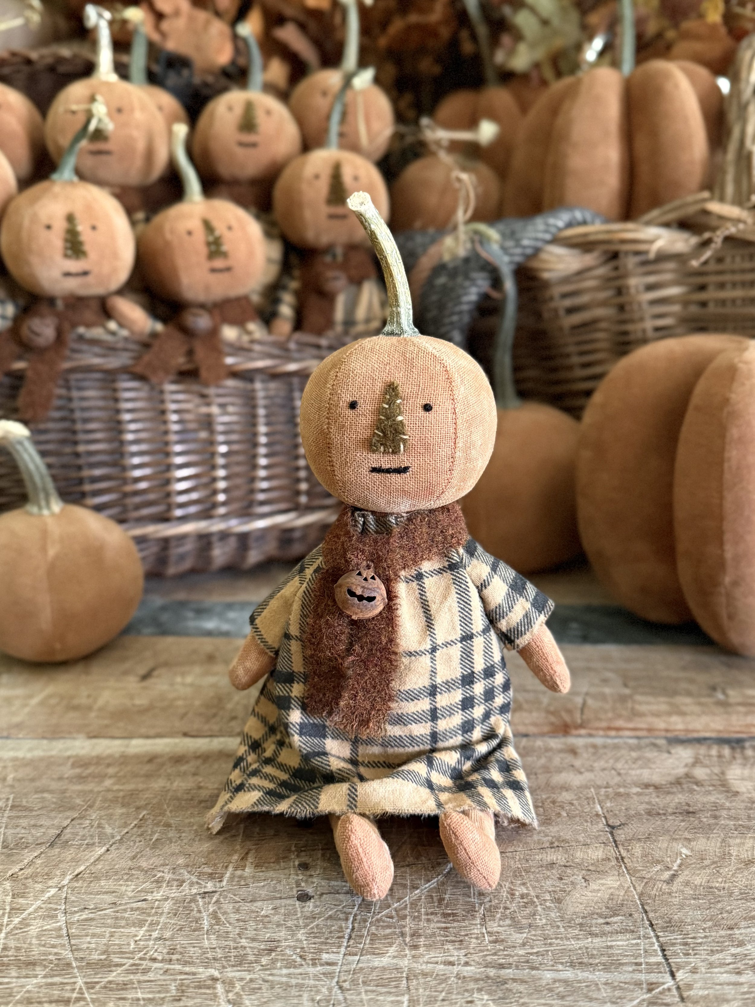 A handmade pumpkin doll with a fabric face featuring a stitched nose and mouth, dressed in a checkered dress, sitting on a wooden surface with a collection of similar pumpkin dolls and pumpkins in the background.