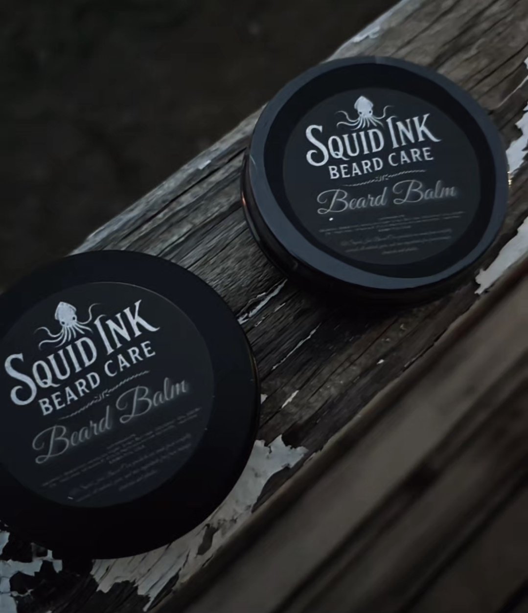 Two round black containers of Squid Ink Beard Care Beard Balm placed on a weathered wooden surface.
