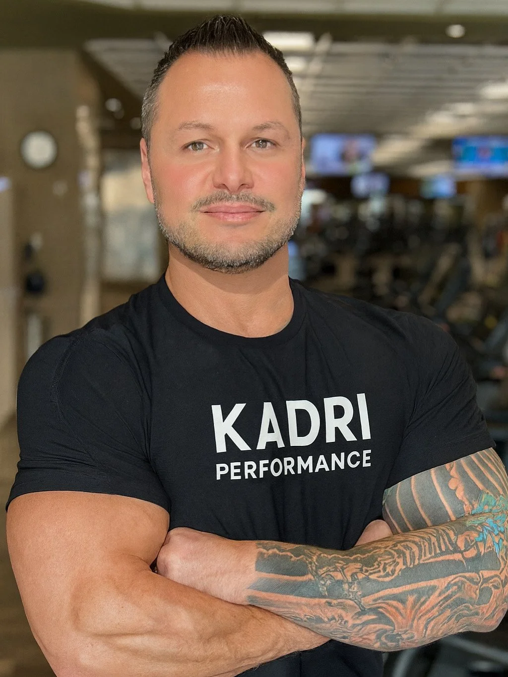 A muscular man with short dark hair and a beard, wearing a black T-shirt with 'KADRI PERFORMANCE' written on it, standing with arms crossed in a gym.