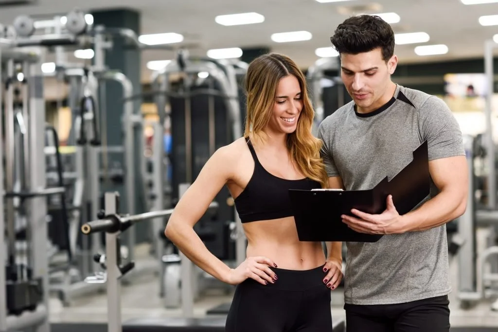 A man and woman in workout clothes standing in a gym, looking at a clipboard together, smiling, with gym equipment in the background.