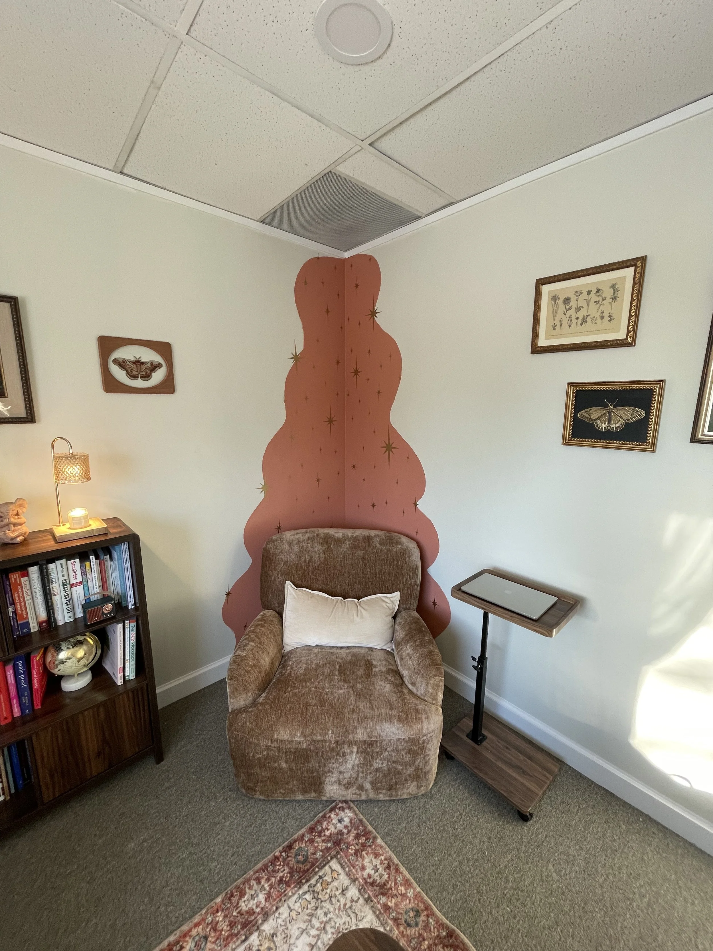 A cozy corner with a brown velvet armchair in front of a pink wall with gold star designs, surrounded by framed botanical and insect artwork, a bookshelf with books, a small table with a tray, and a patterned rug.