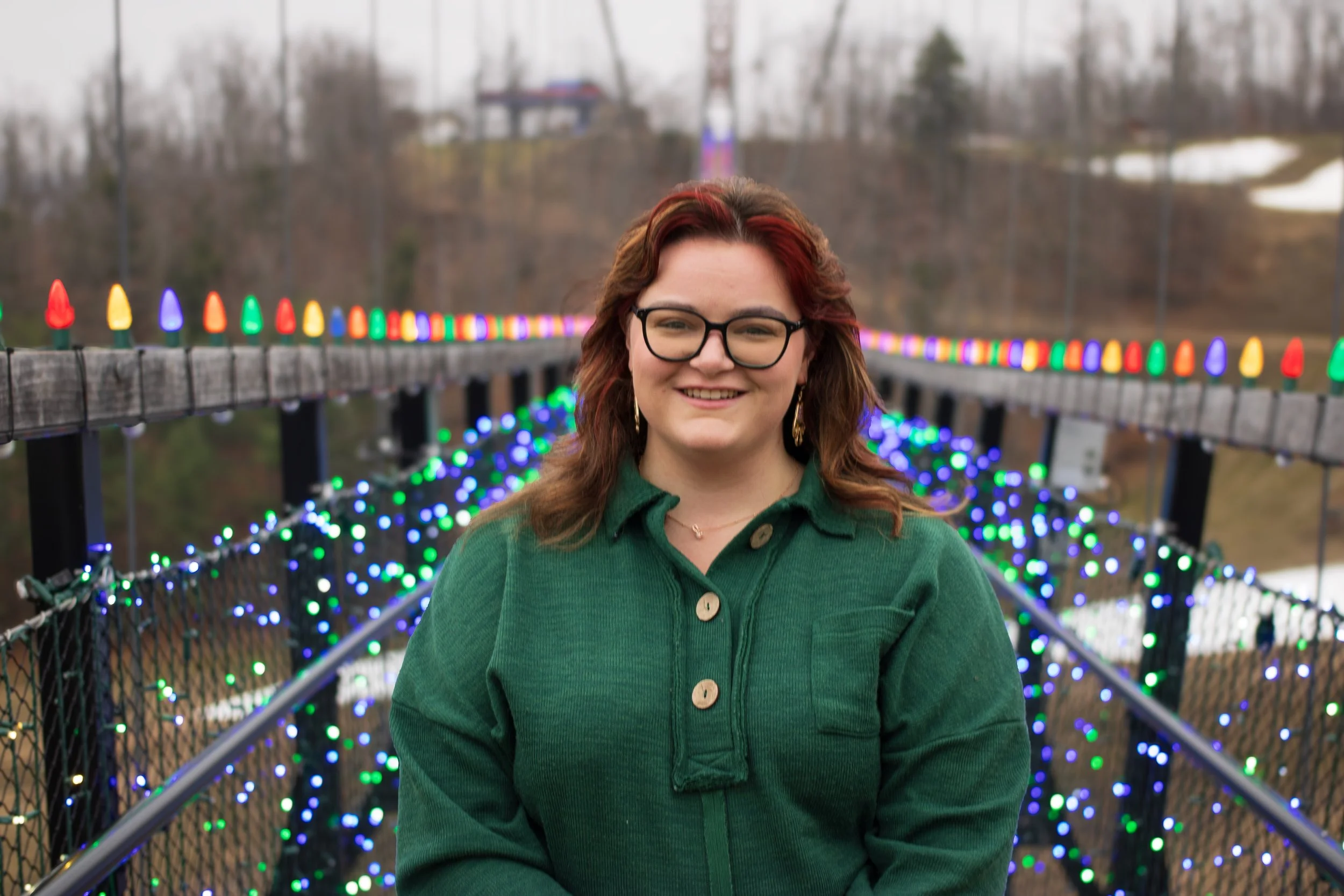A woman with glasses, red-brown hair, and earrings standing on a bridge decorated with colorful holiday lights, with a wooded area in the background.