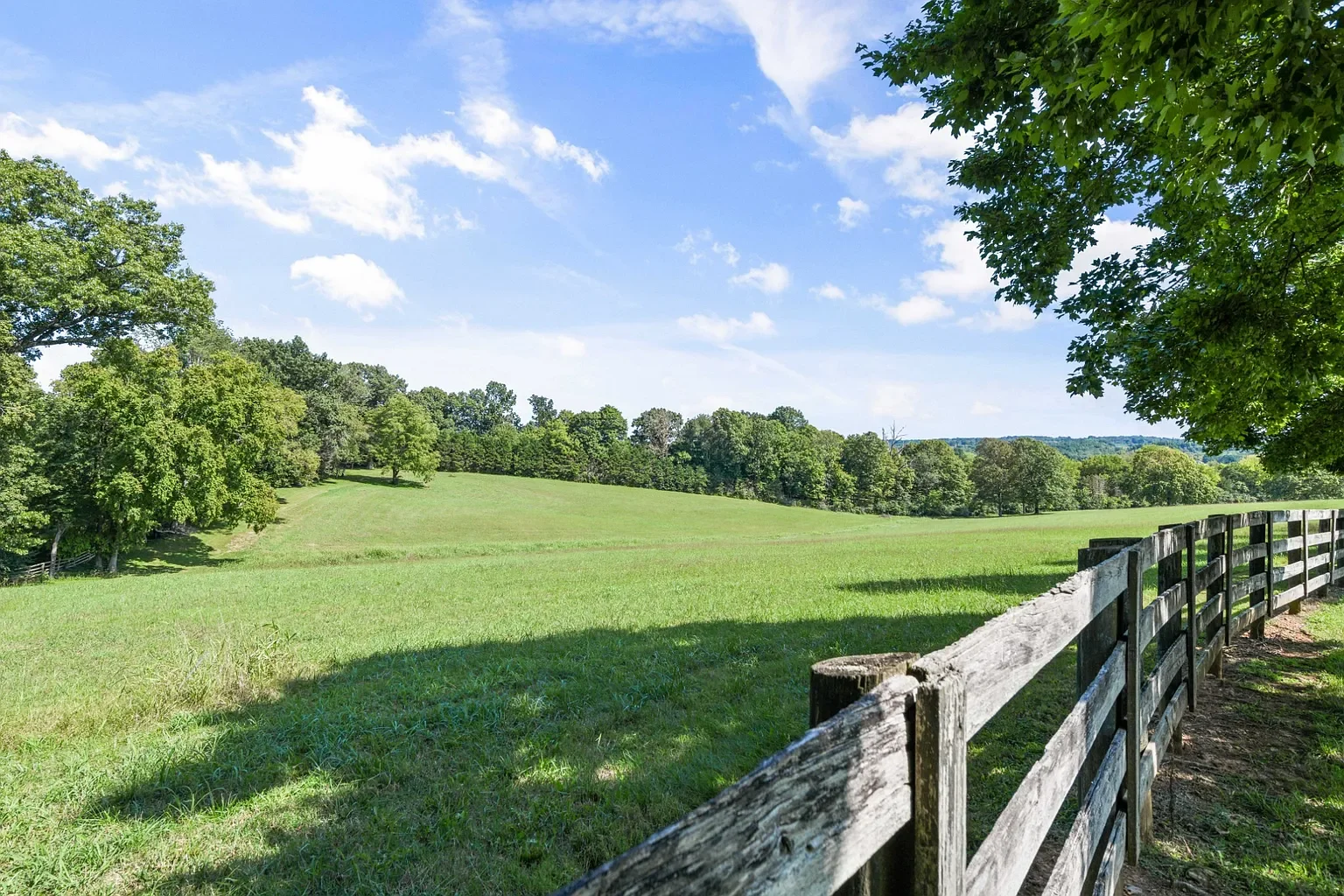 Green rolling hills with scattered trees and a fork in the distance, clear blue sky with a few clouds, and a wooden fence along the right side of the image.
