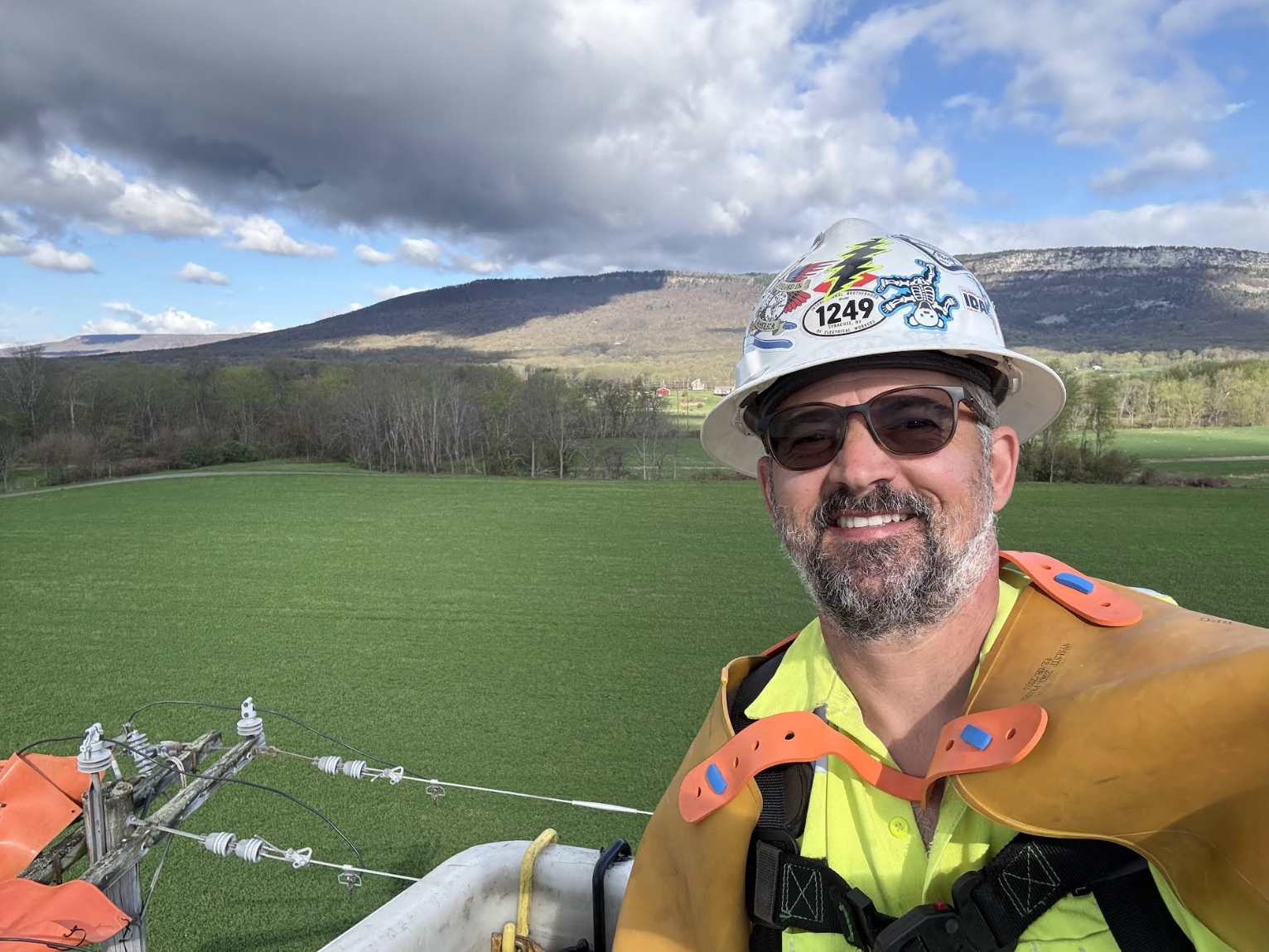 Eric, Owner of Wave That Flag, smiling while in a lineman bucket with a view of the Shawangunk Mountain Ridge and rolling landscape hills behind him