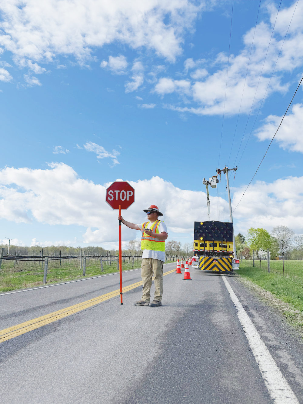 Work zone flagger holding a stop sign on a rural road with utility trucks and orange cones, under a partly cloudy sky.