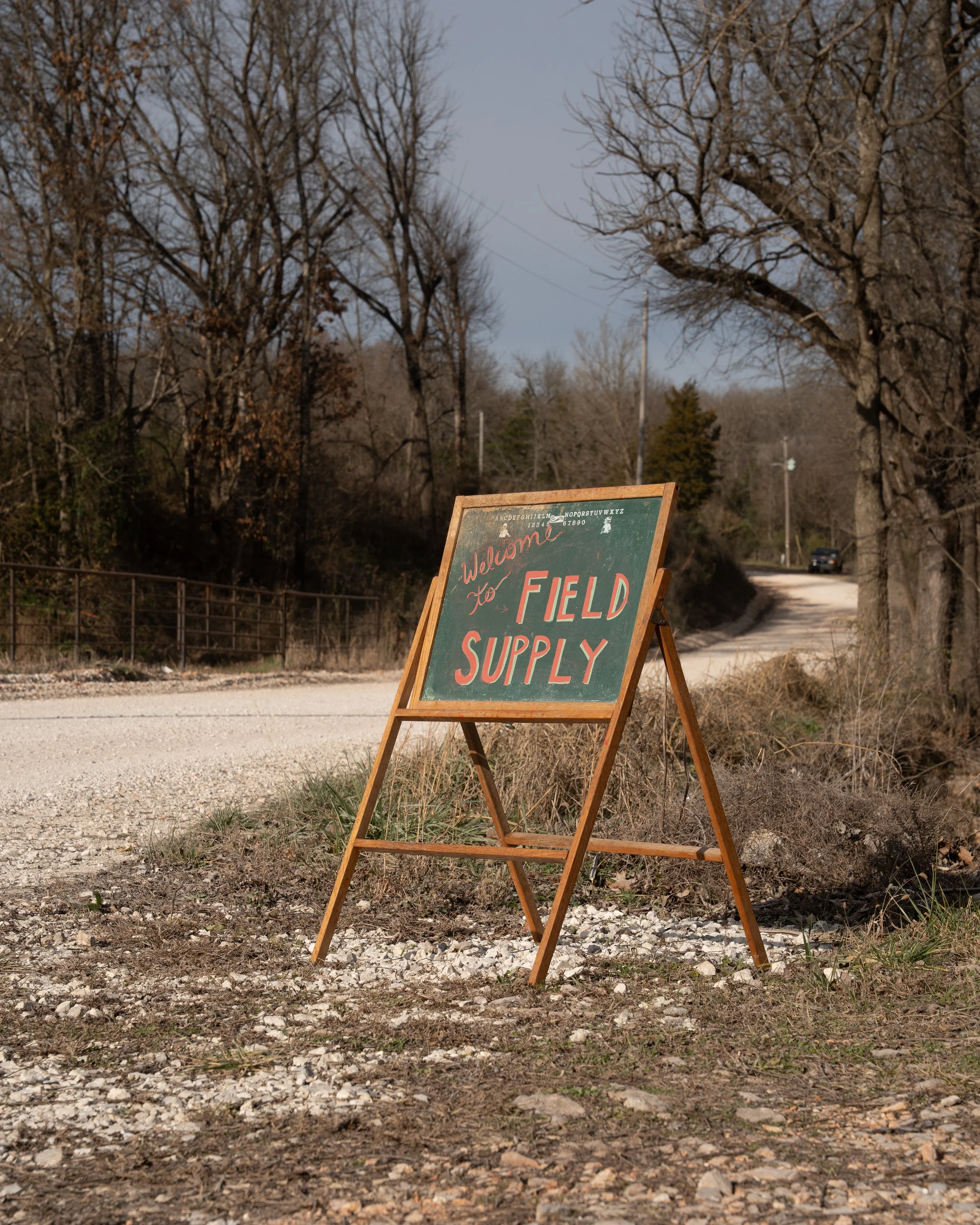 A green and red signboard on a wooden stand that reads 'Welcome to Field Supply' placed on a gravel roadside with leafless trees and a winding road in the background.