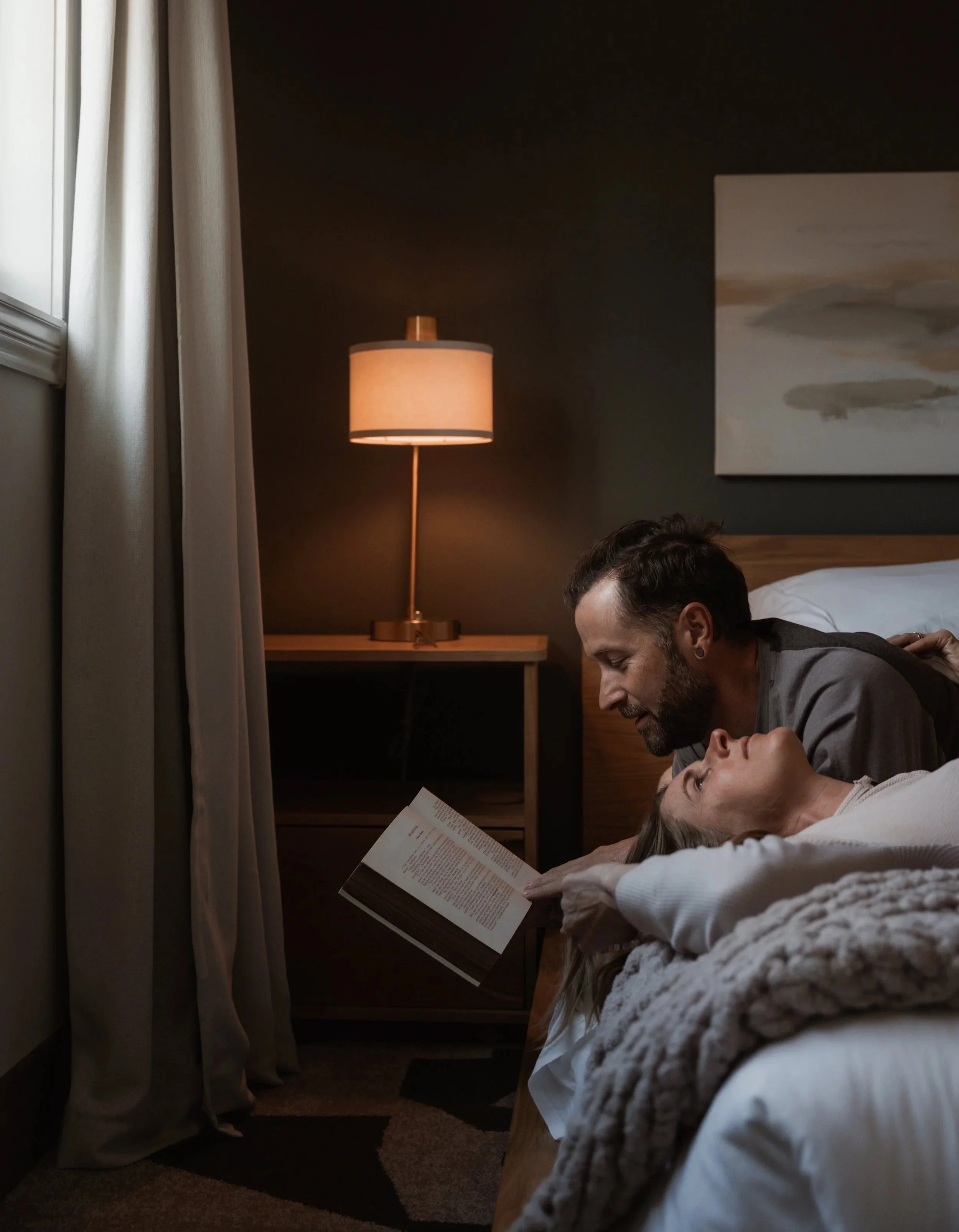 A couple lying in bed, reading a book together in a cozy bedroom with a lamp on a nightstand and curtains.
