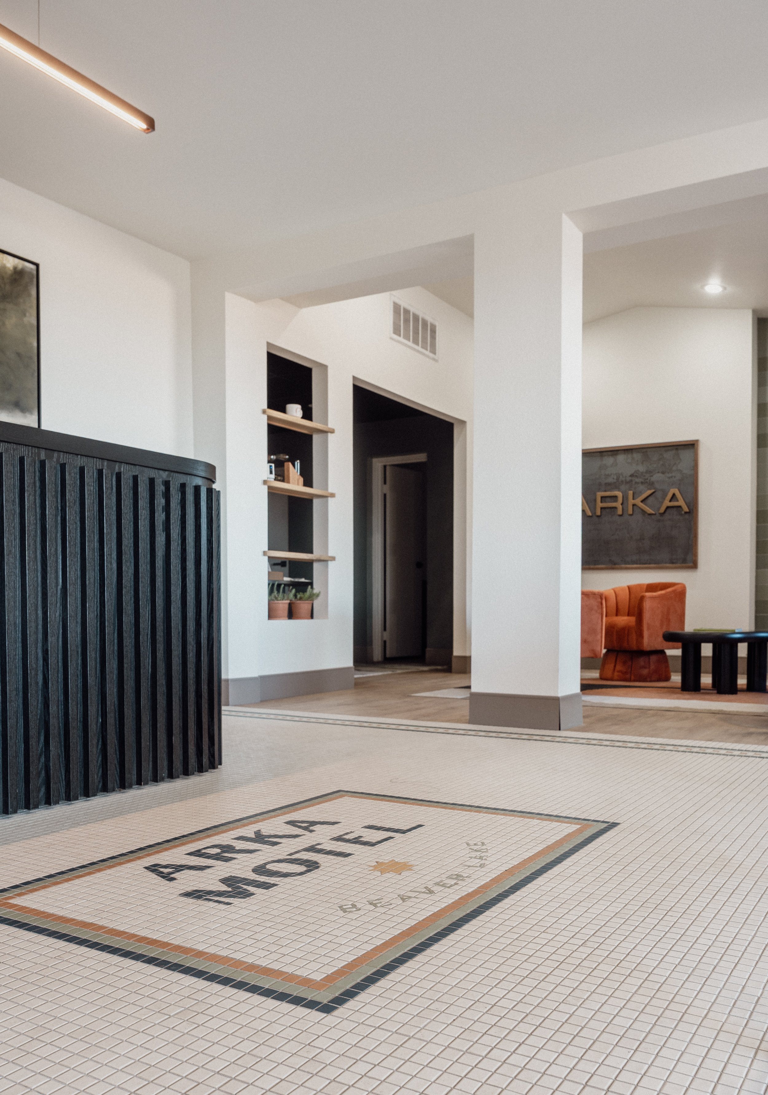 Interior of a modern lobby with a tiled floor, a black slatted counter, white walls, and a seating area with an orange sofa and black coffee table. There is wall art with the word 'ARKA' on it.