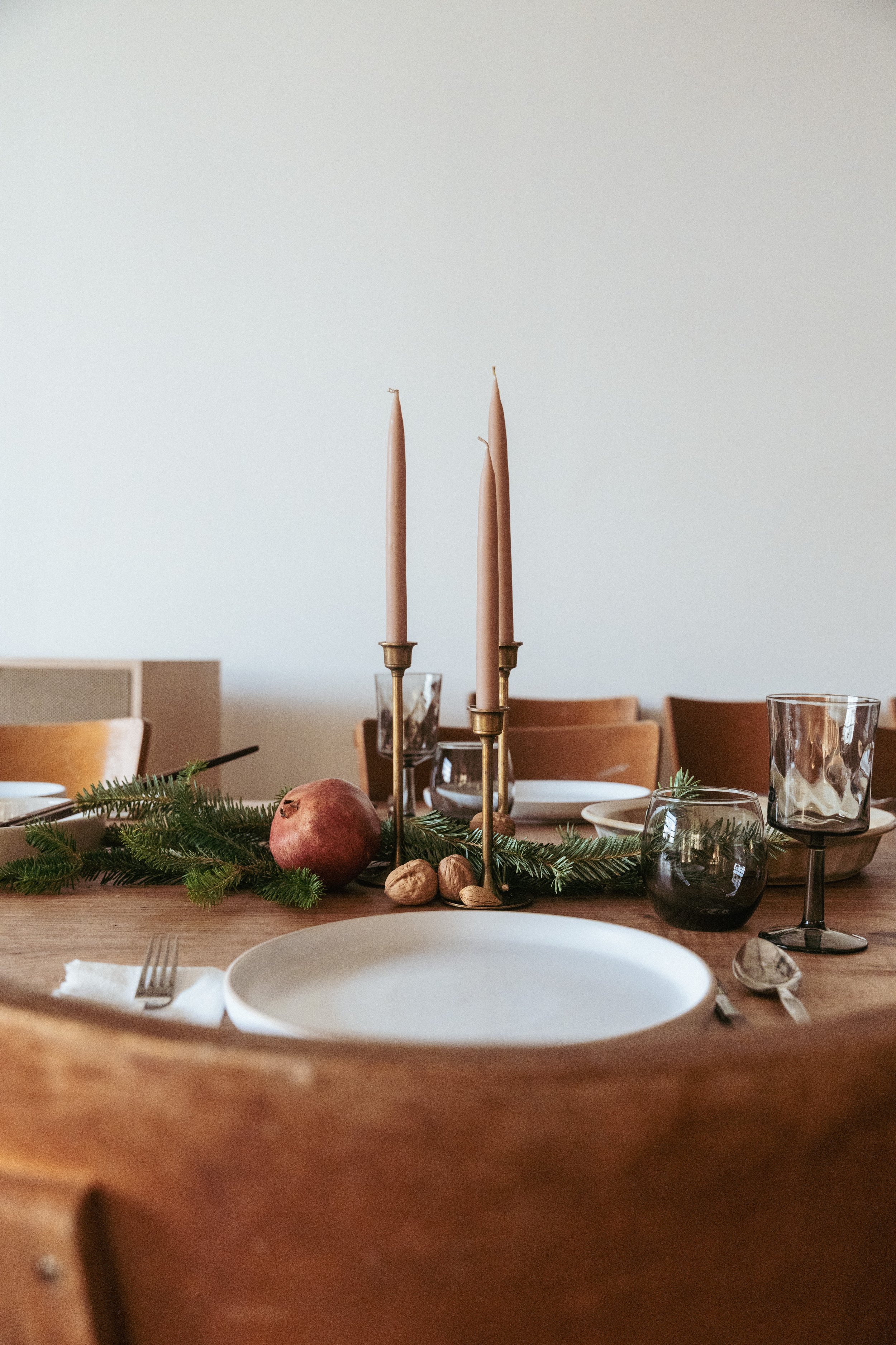 A wooden dining table set for a meal with a white plate, glassware, and silverware, decorated with green pine branches, pink candles in brass holders, a pomegranate, and nuts.