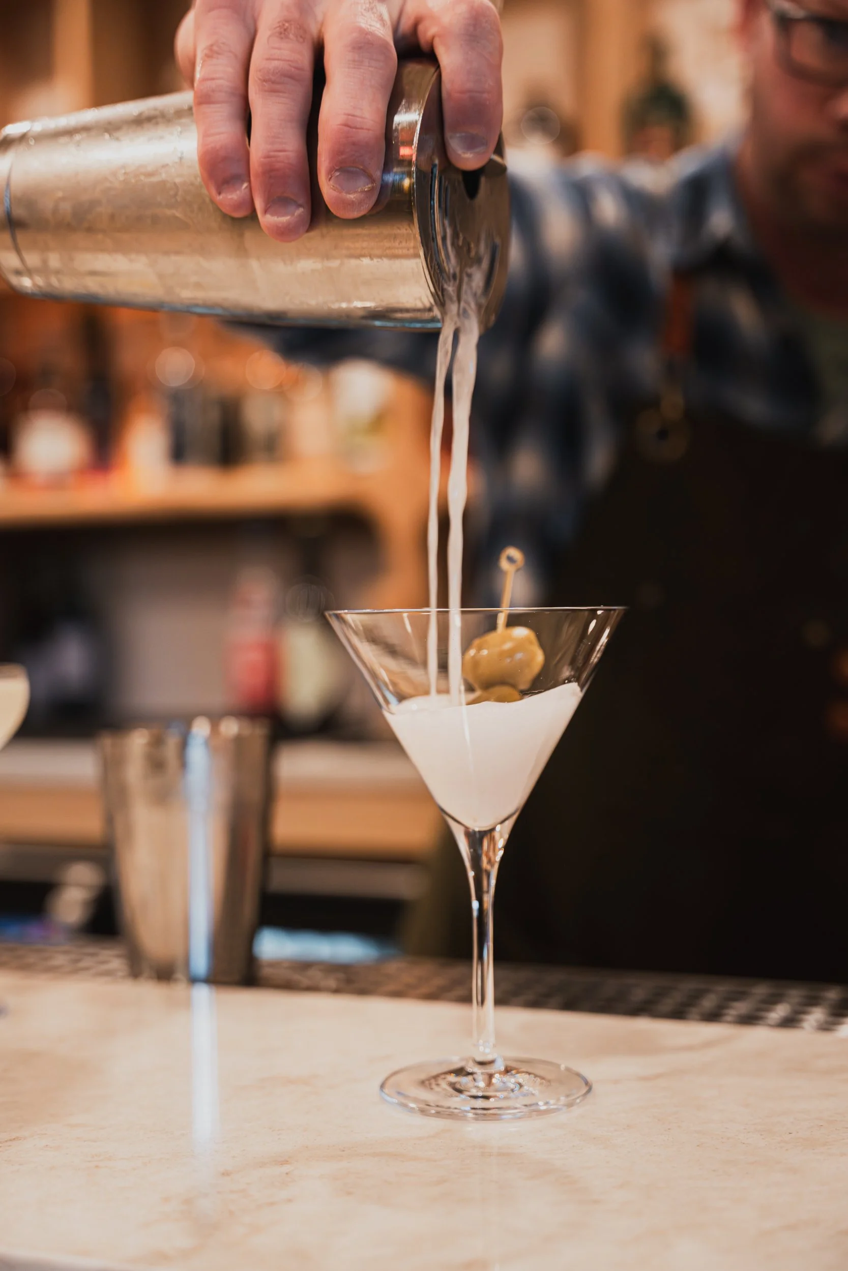 A bartender pouring a cocktail from a shaker into a martini glass with an olive in it.