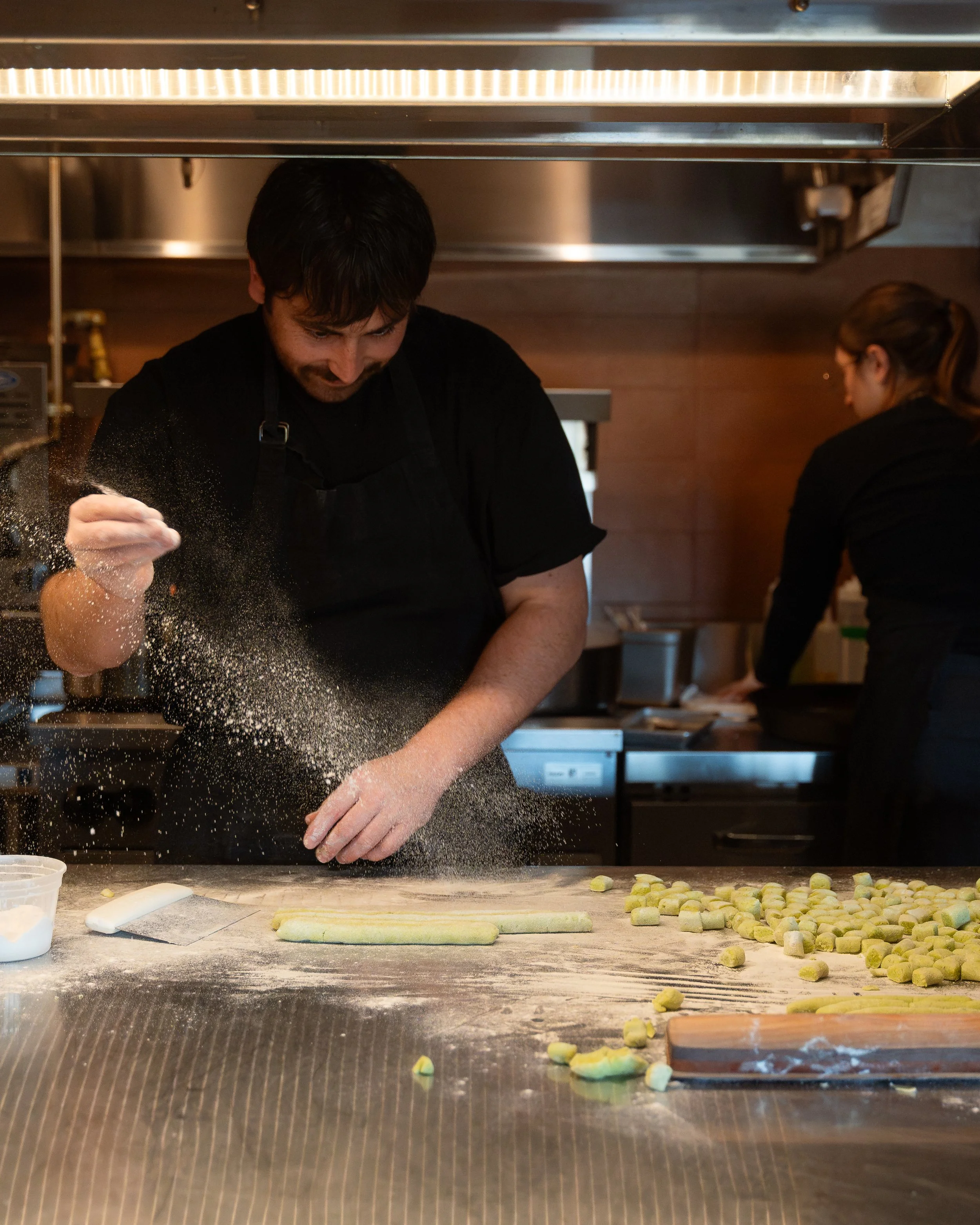 A baker dusts flour onto dough on a kitchen counter, surrounded by small dough pieces, with another person working in the background.