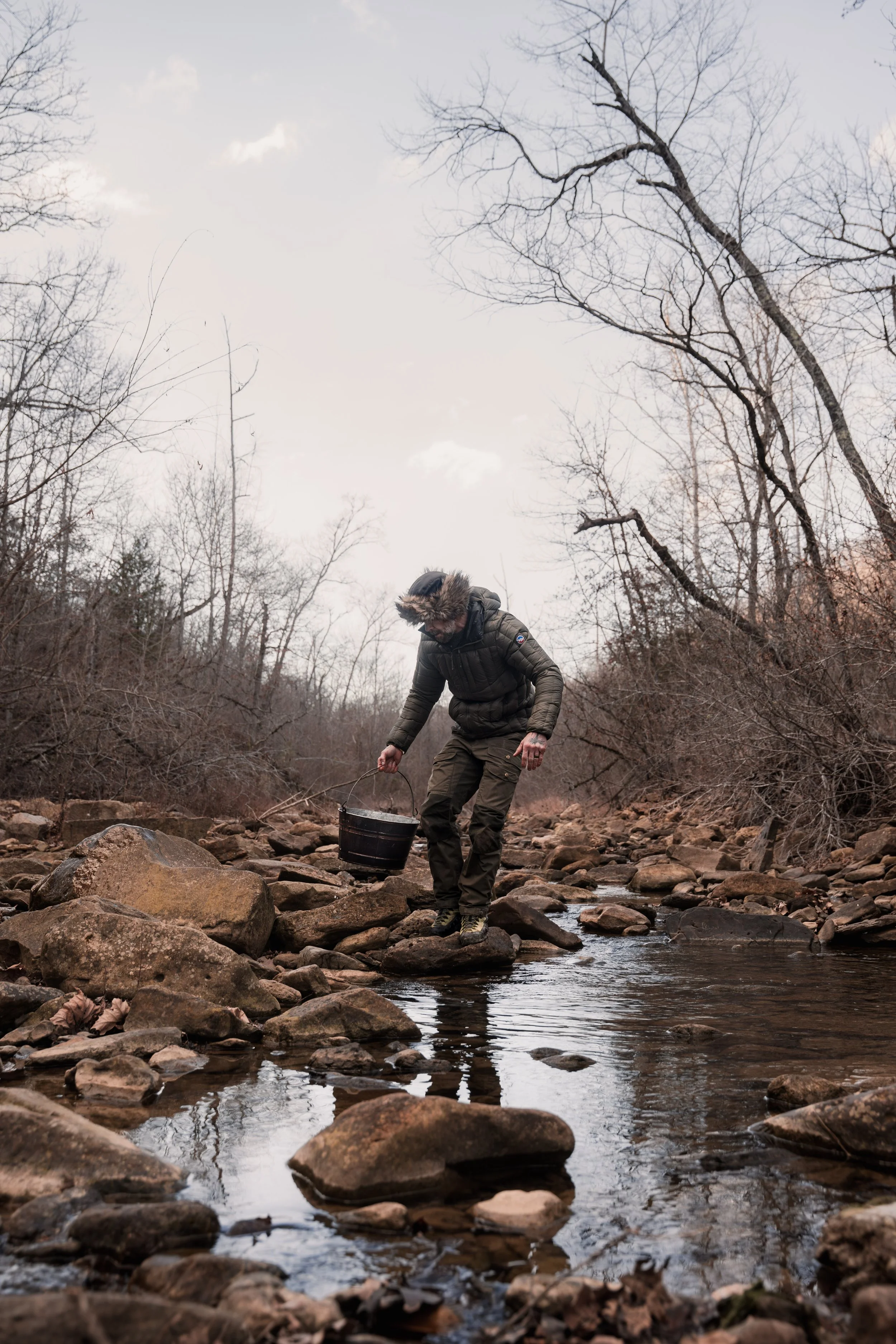 A person in outdoor winter clothing standing on rocks in a creek, holding a bucket, with leafless trees in the background.