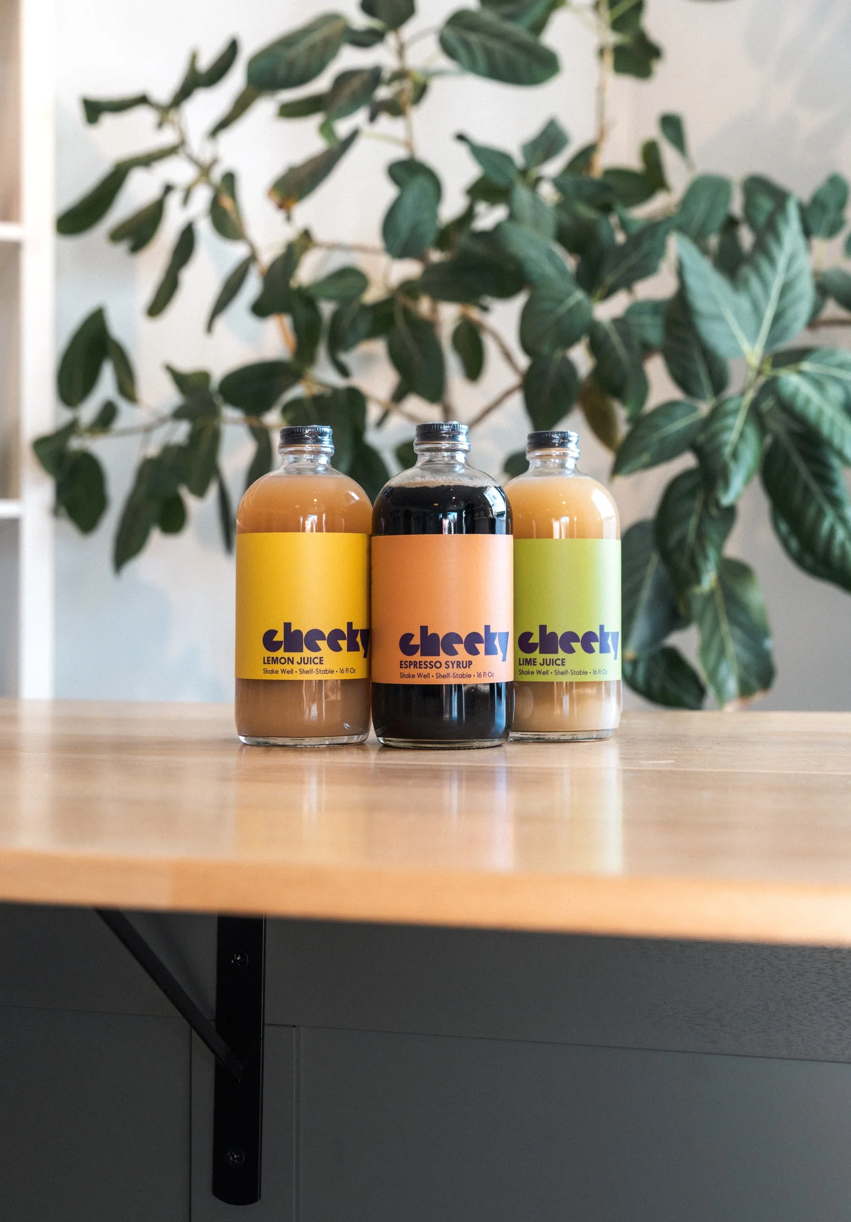 Three bottles of Cheeky juice on a wooden table with a leafy green plant in the background.