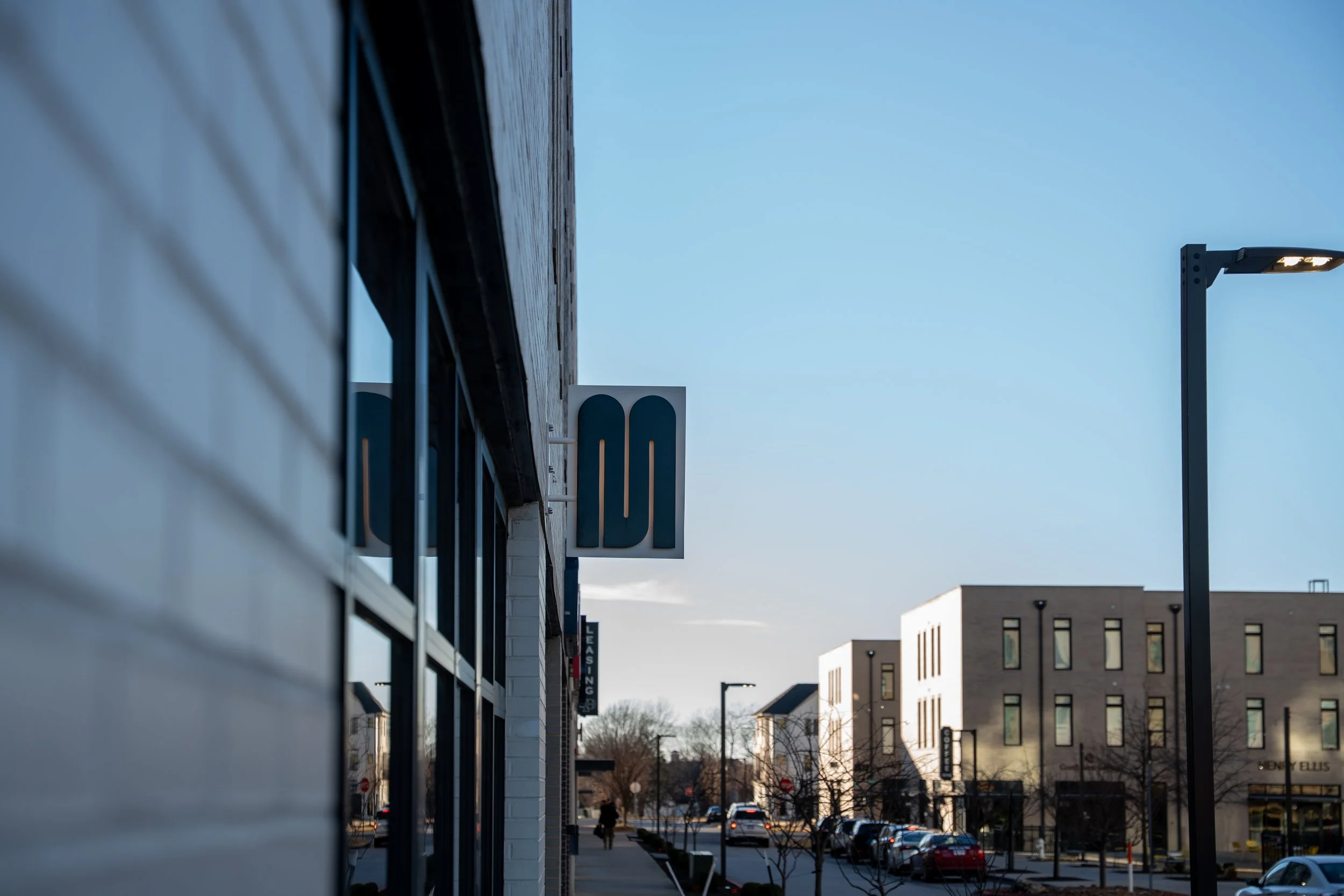 Street view in a modern commercial area with white buildings, parked cars, streetlights, and a blue sky.