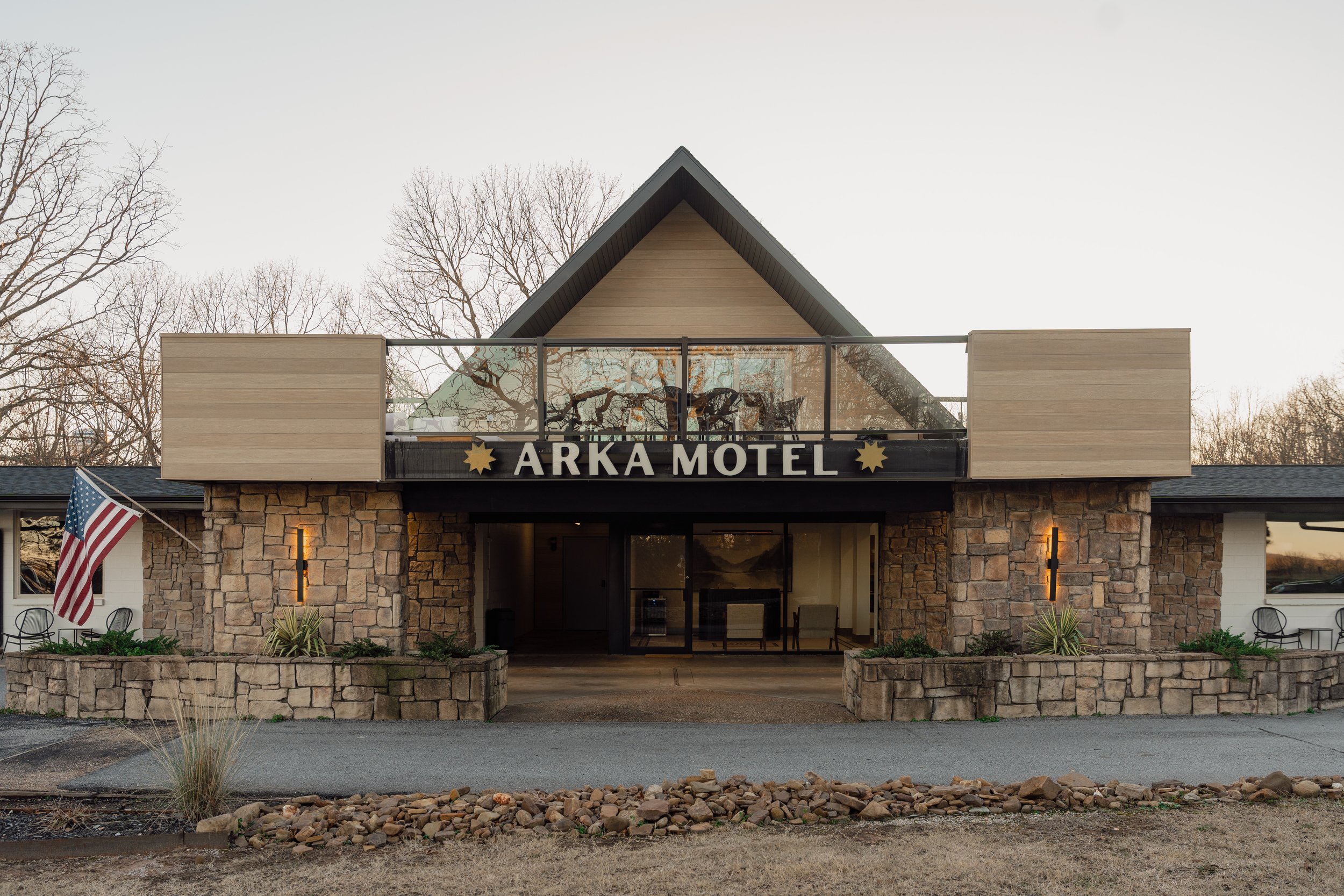 Front view of the Arka Motel with brick and stone exterior, outdoor seating, and an American flag, during sunset or late afternoon.