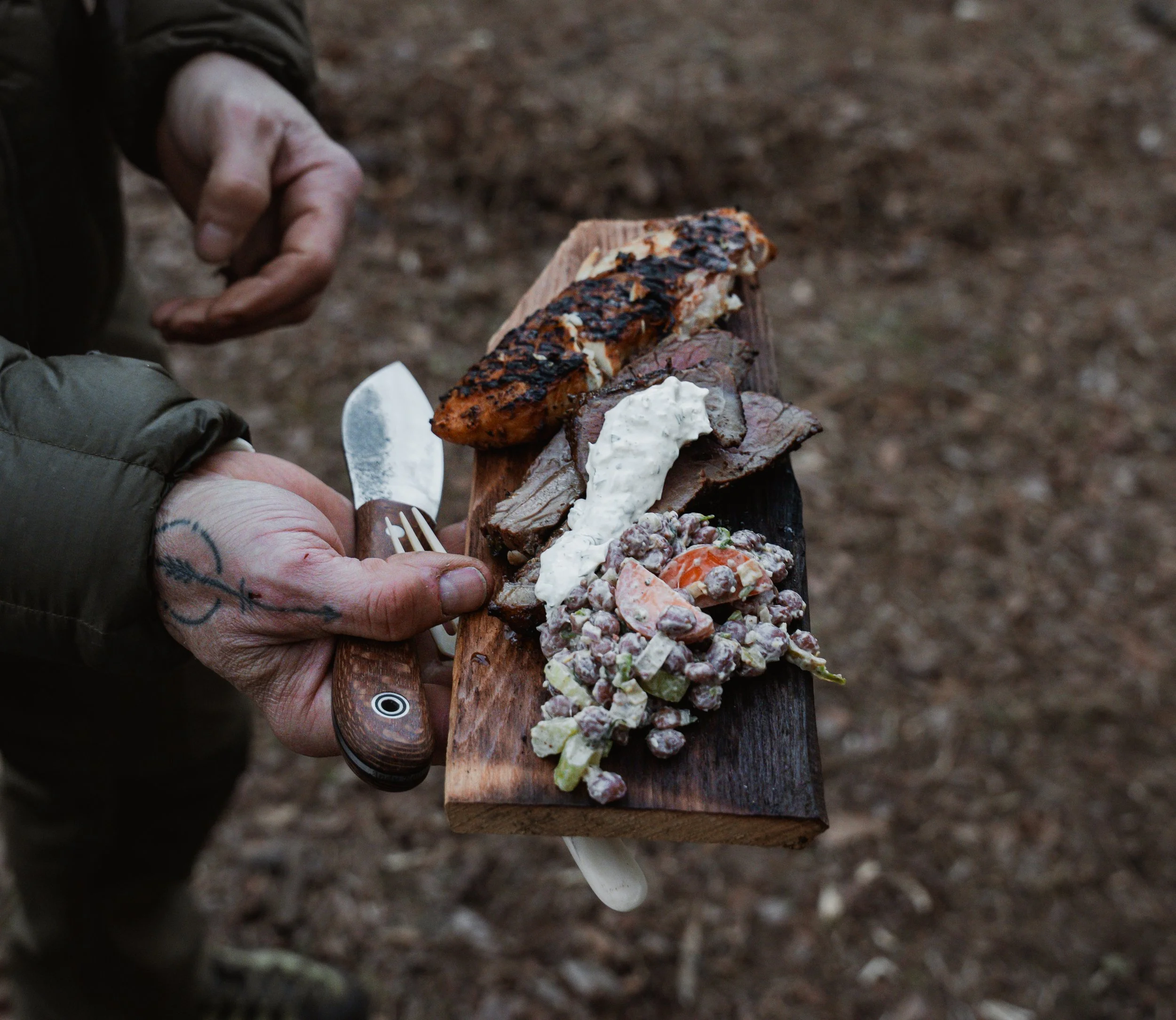 Person holding a wooden board with grilled chicken, slices of beef, a dollop of white sauce, and a salad with beans and vegetables. The person is using a fork and a knife, and the background appears to be an outdoor dirt ground.
