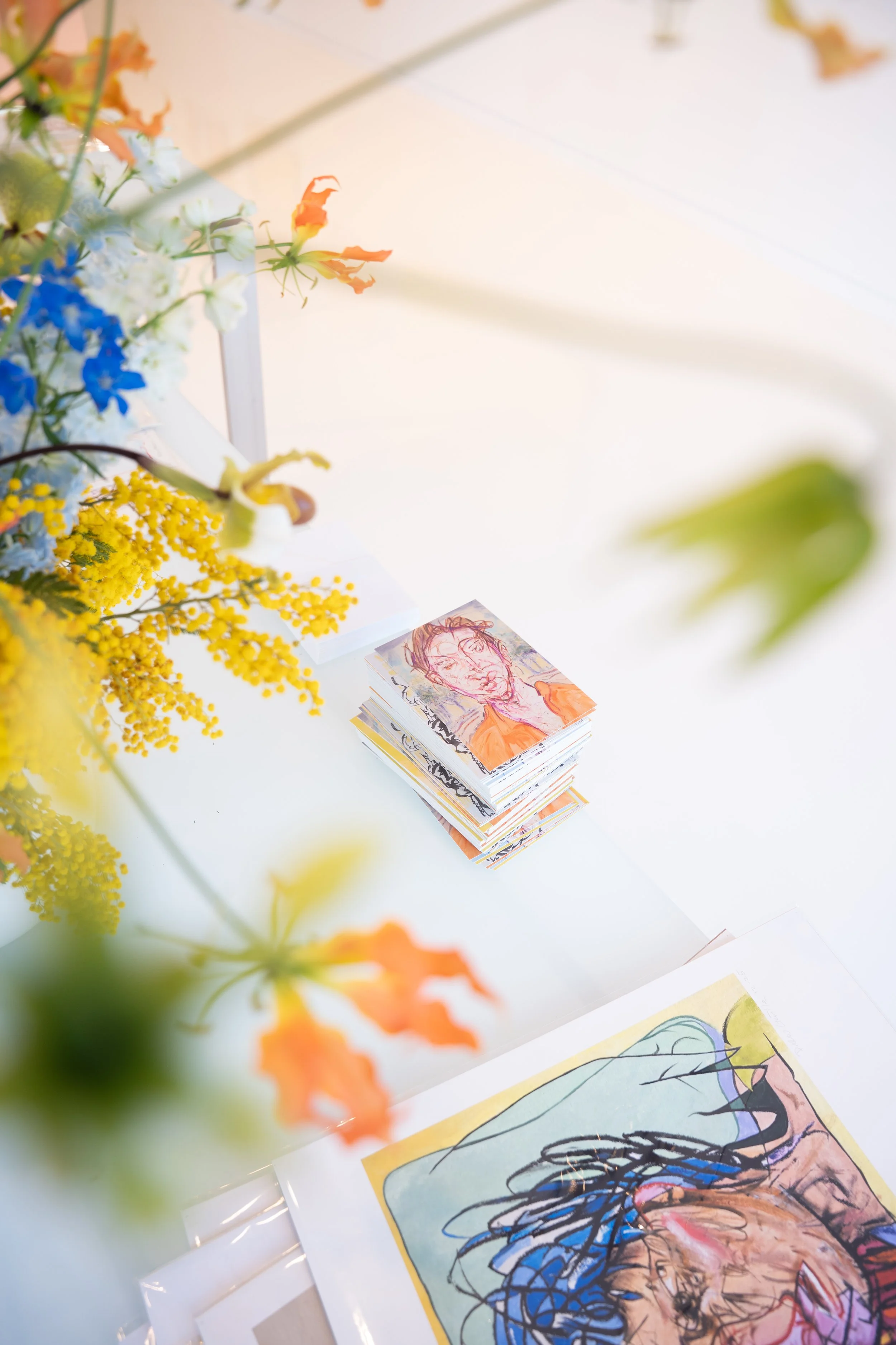 A white table with a colorful, abstract portrait drawing, a stack of smaller artworks with a portrait, and a vase of variously colored flowers including yellow, blue, white, and orange.