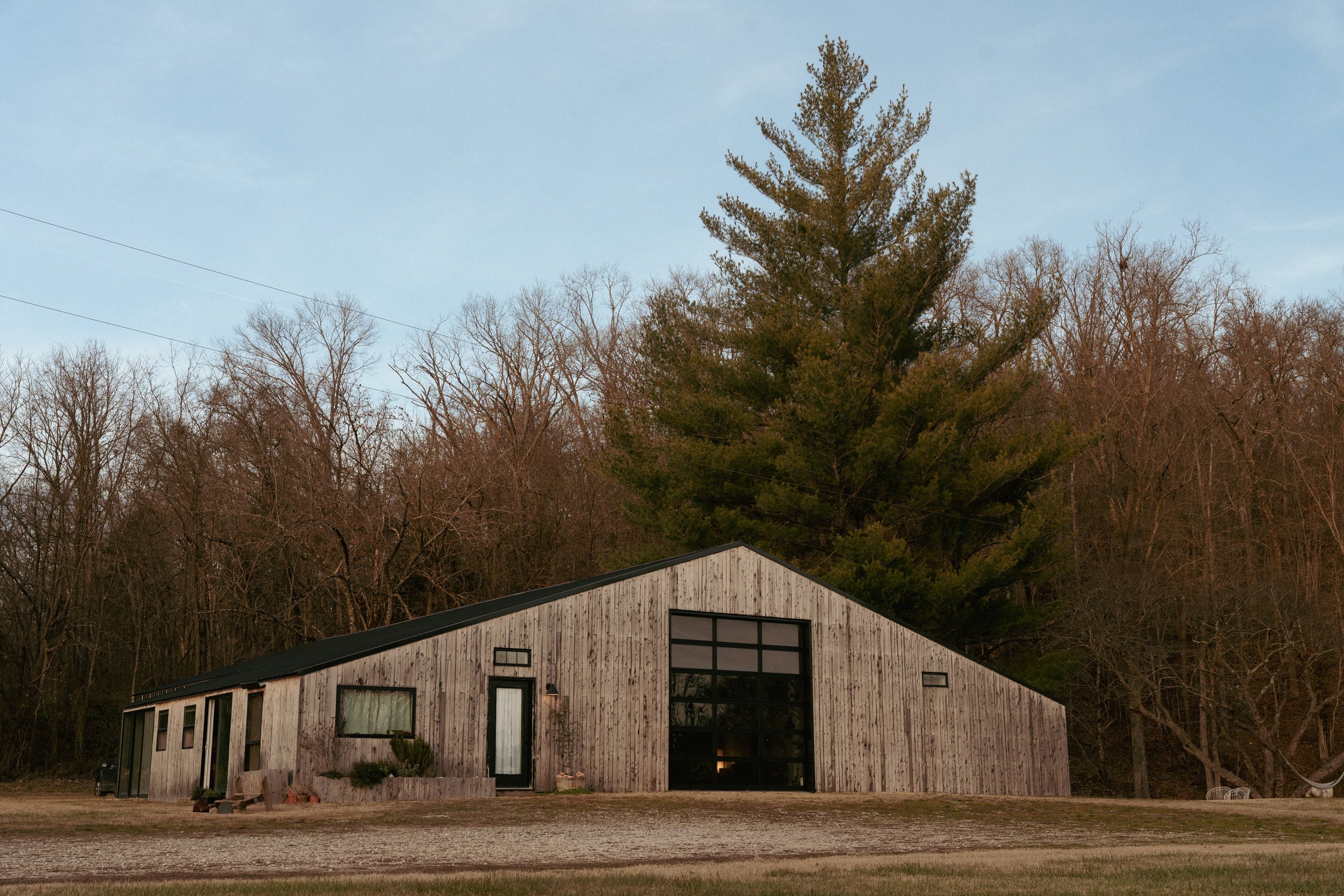 A rustic wooden barn with large glass garage door, surrounded by leafless trees, with a large pine tree in the background under a partly cloudy sky.