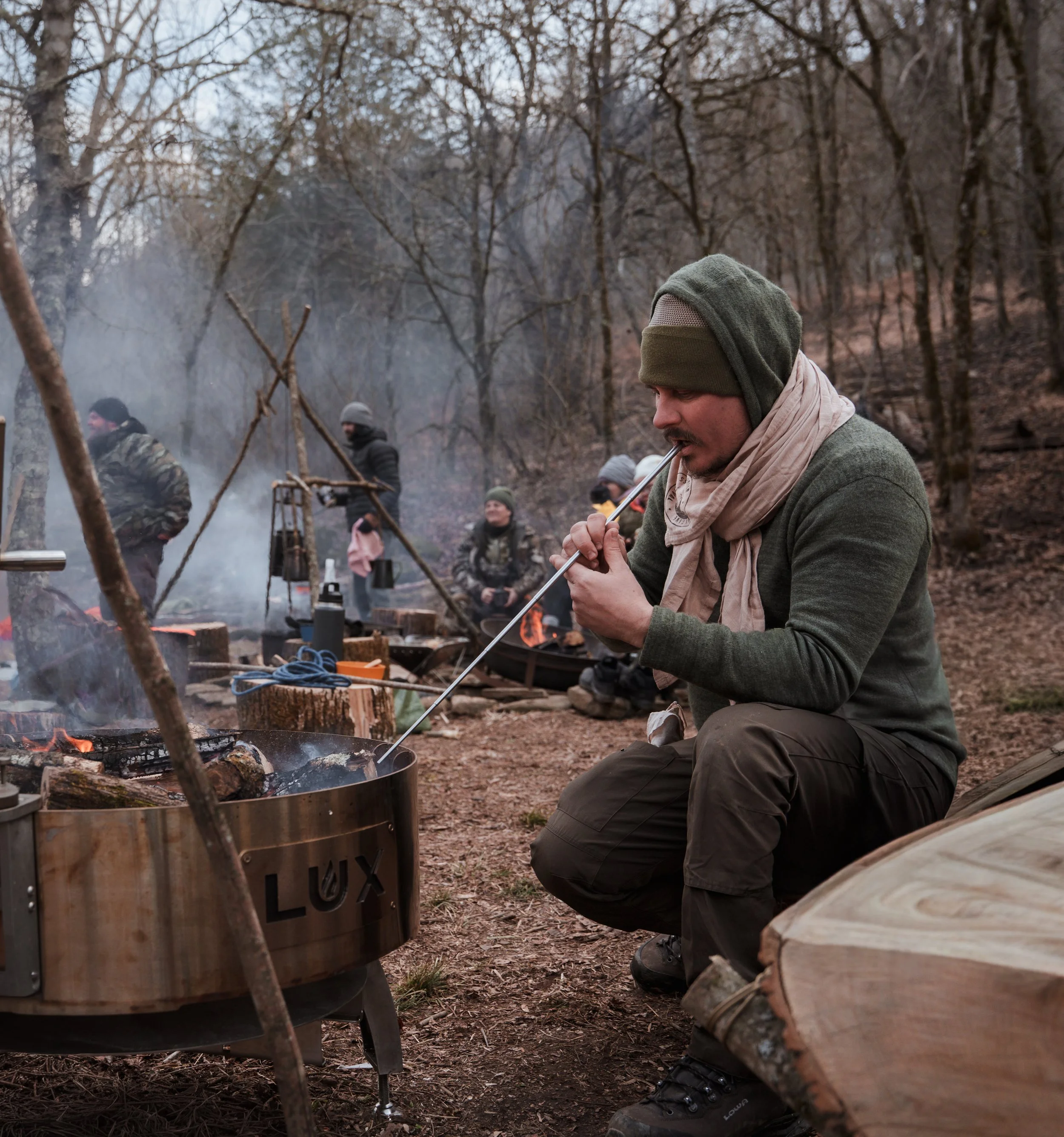 A man sitting outdoors around a campfire, using a long stick to cook or prepare food. Other people are gathered around in the background in a wooded area during what appears to be late fall or winter.