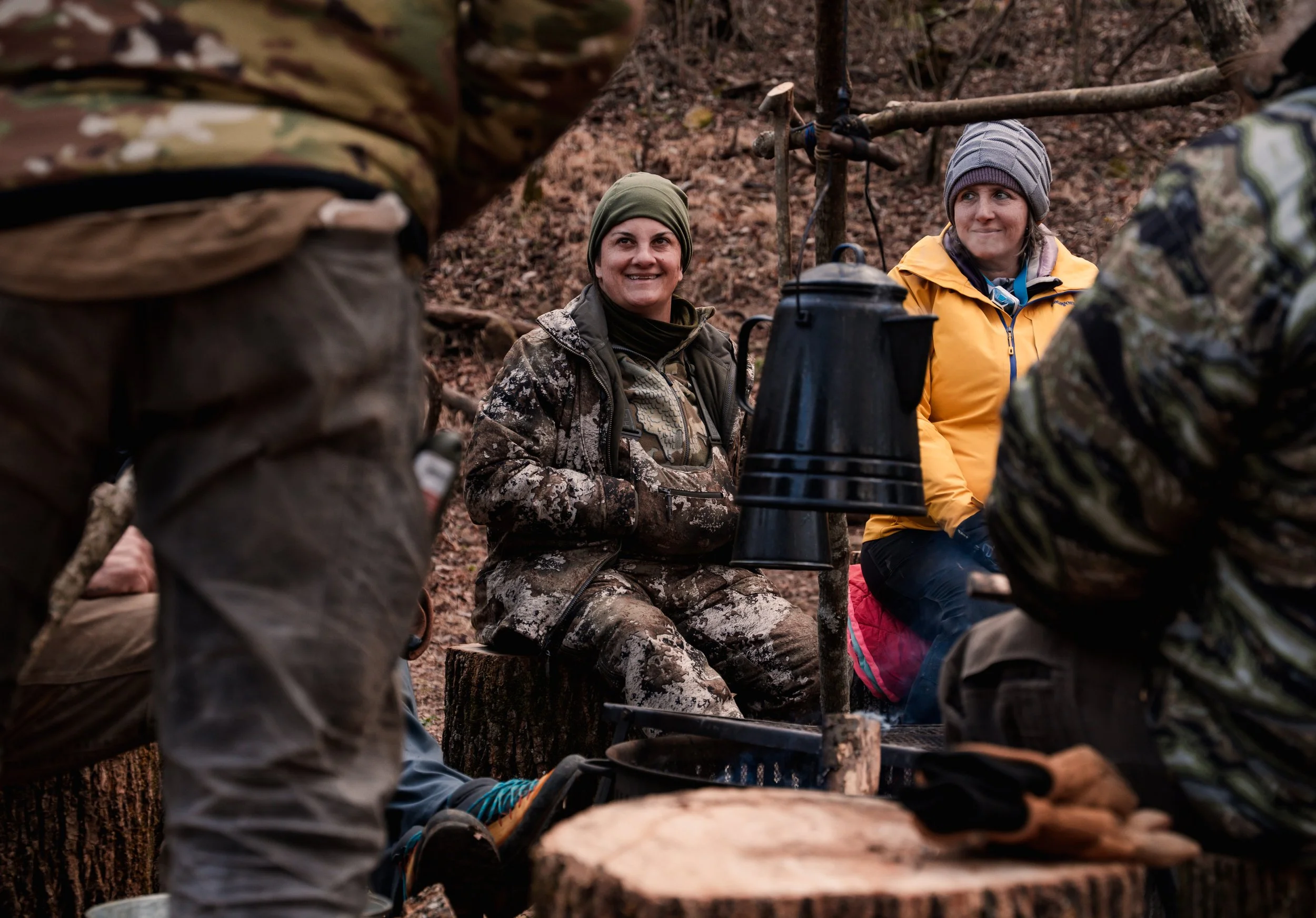 Group of people dressed in outdoor clothing, sitting in a forest, having a discussion around a makeshift table with a black coffee pot hanging from a branch.