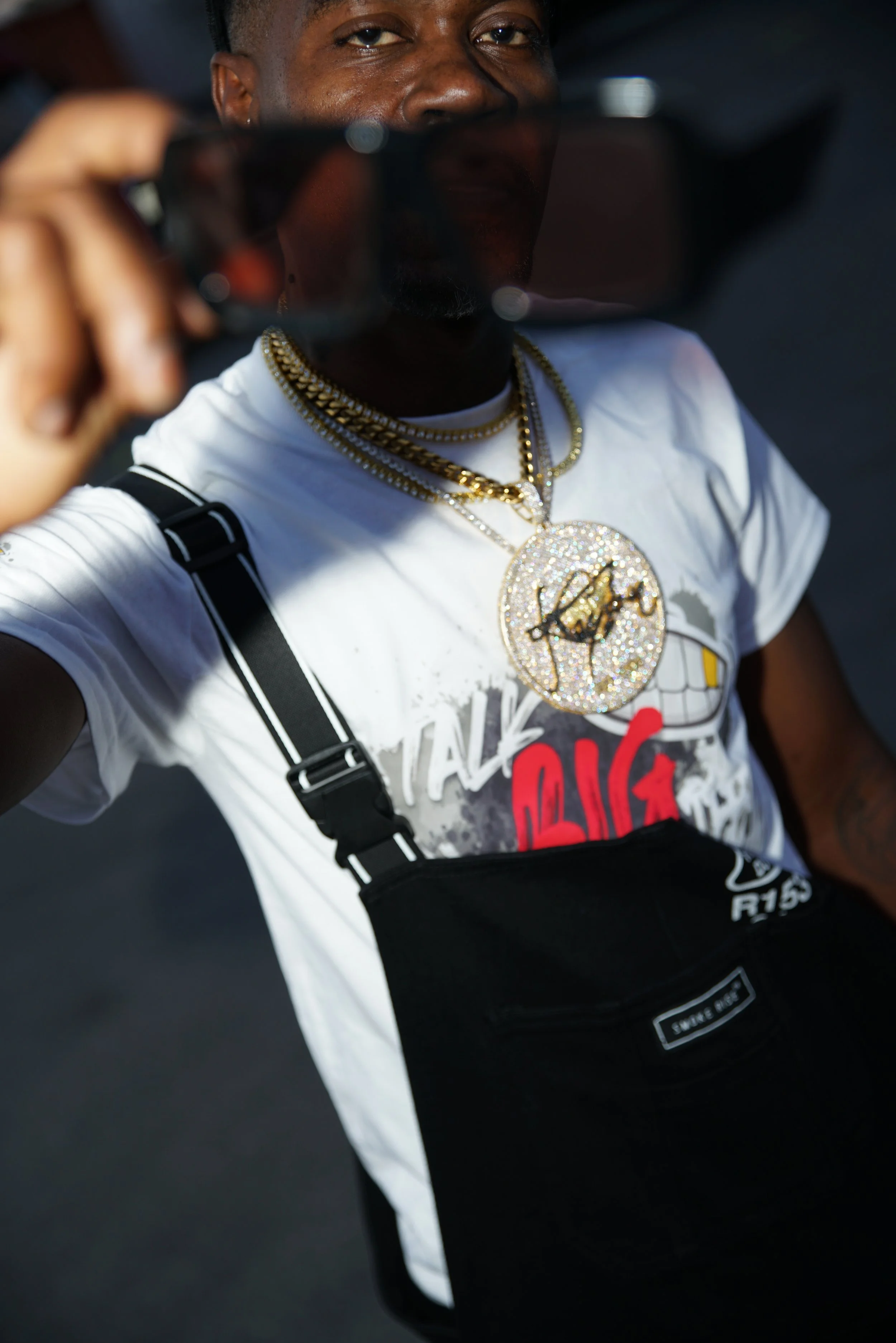 A person wearing multiple gold chains, some with large pendants, a white graphic t-shirt, black suspenders, and carrying a black bag, taking a selfie.