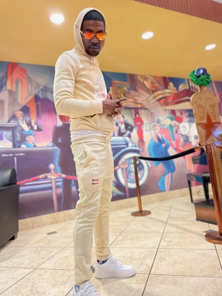 Young man in beige hoodie, matching beige sweatpants, white sneakers, and orange sunglasses standing indoors against colorful mural background, holding a phone.