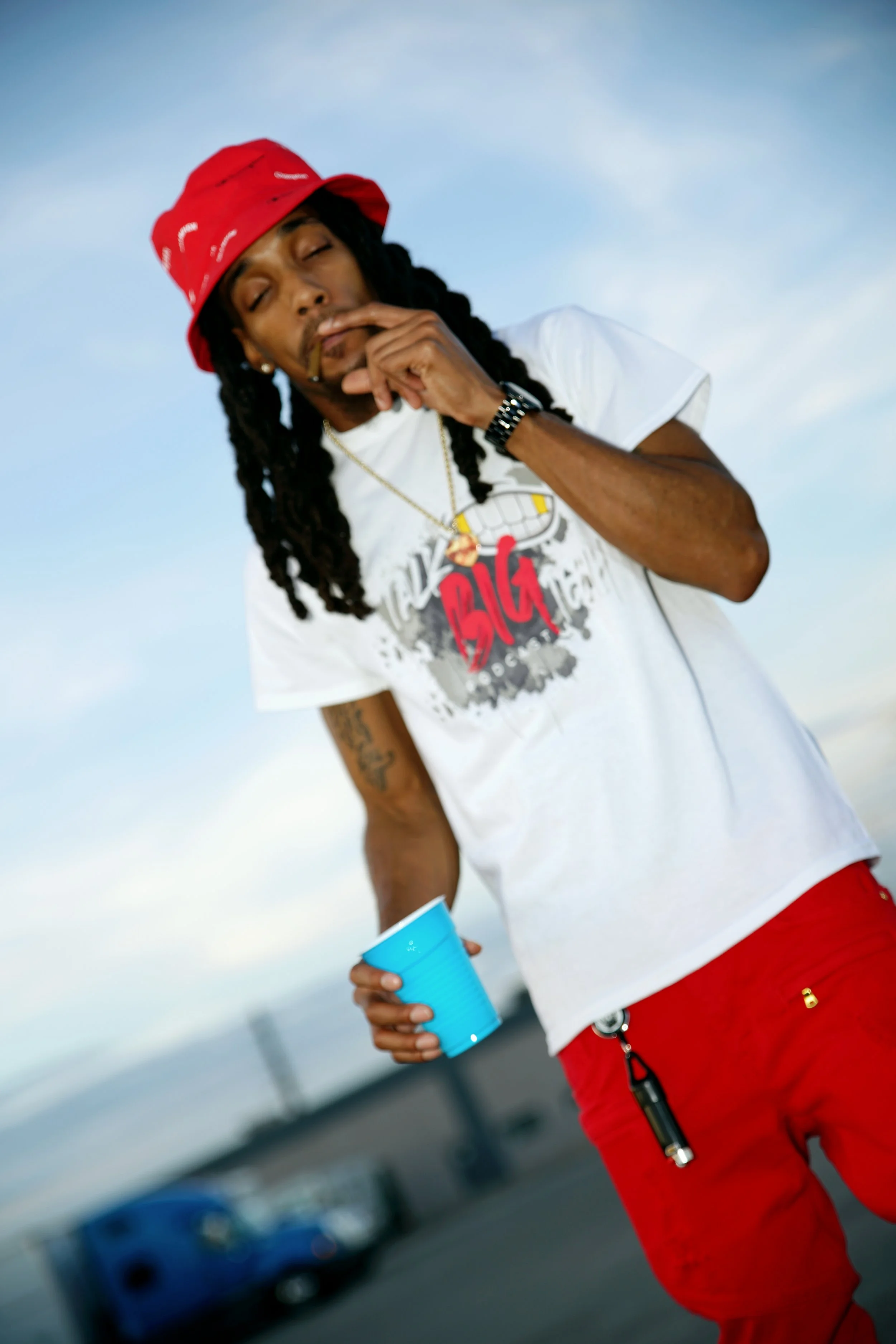 A man with dreadlocks wearing a red bucket hat, a white t-shirt with graphic design, and red pants, holding a blue cup, standing outdoors against a cloudy sky.