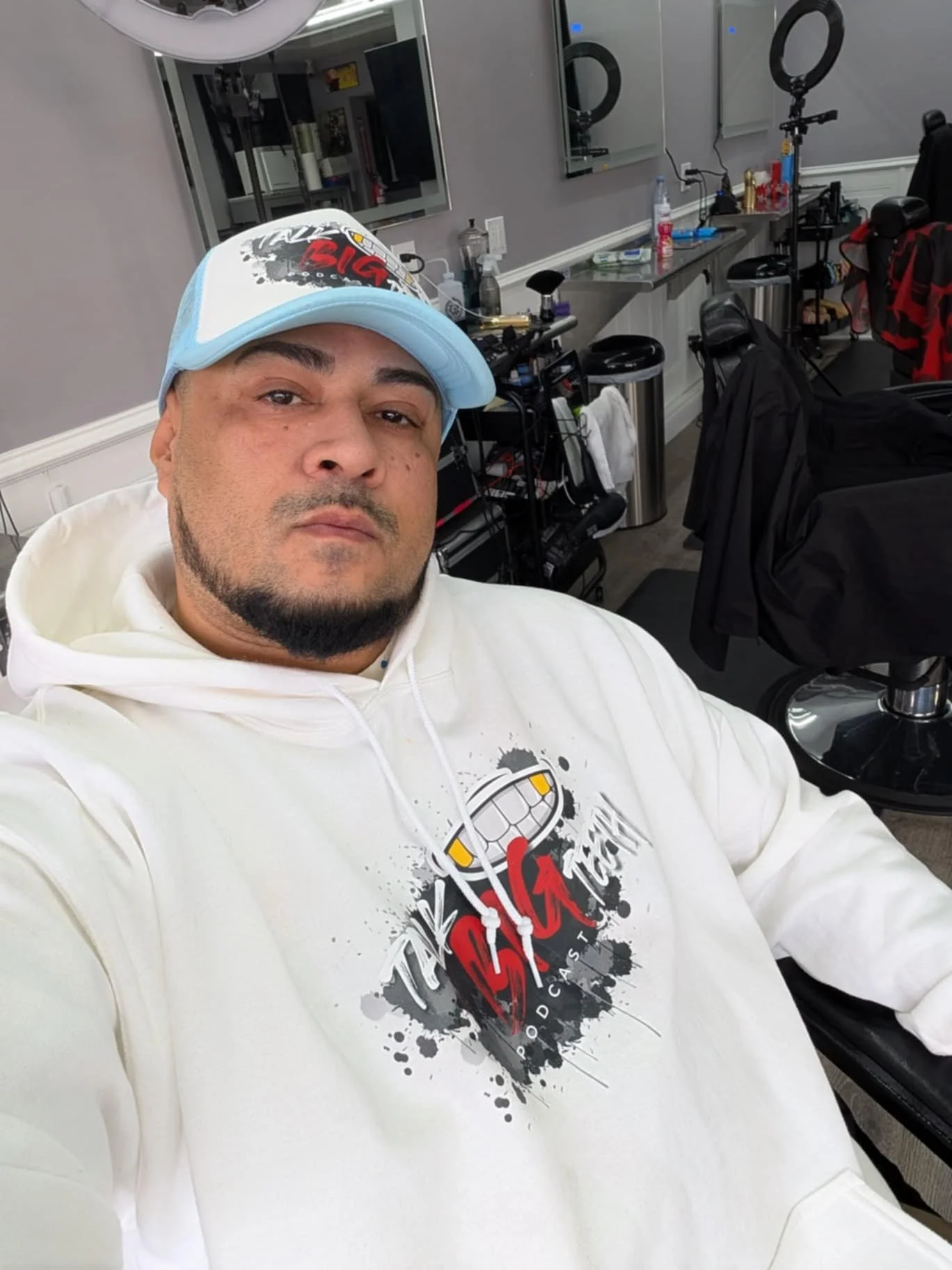 Man taking a selfie in a barbershop, wearing a white hoodie with a logo and a blue cap, with barber chairs and equipment visible in the background.