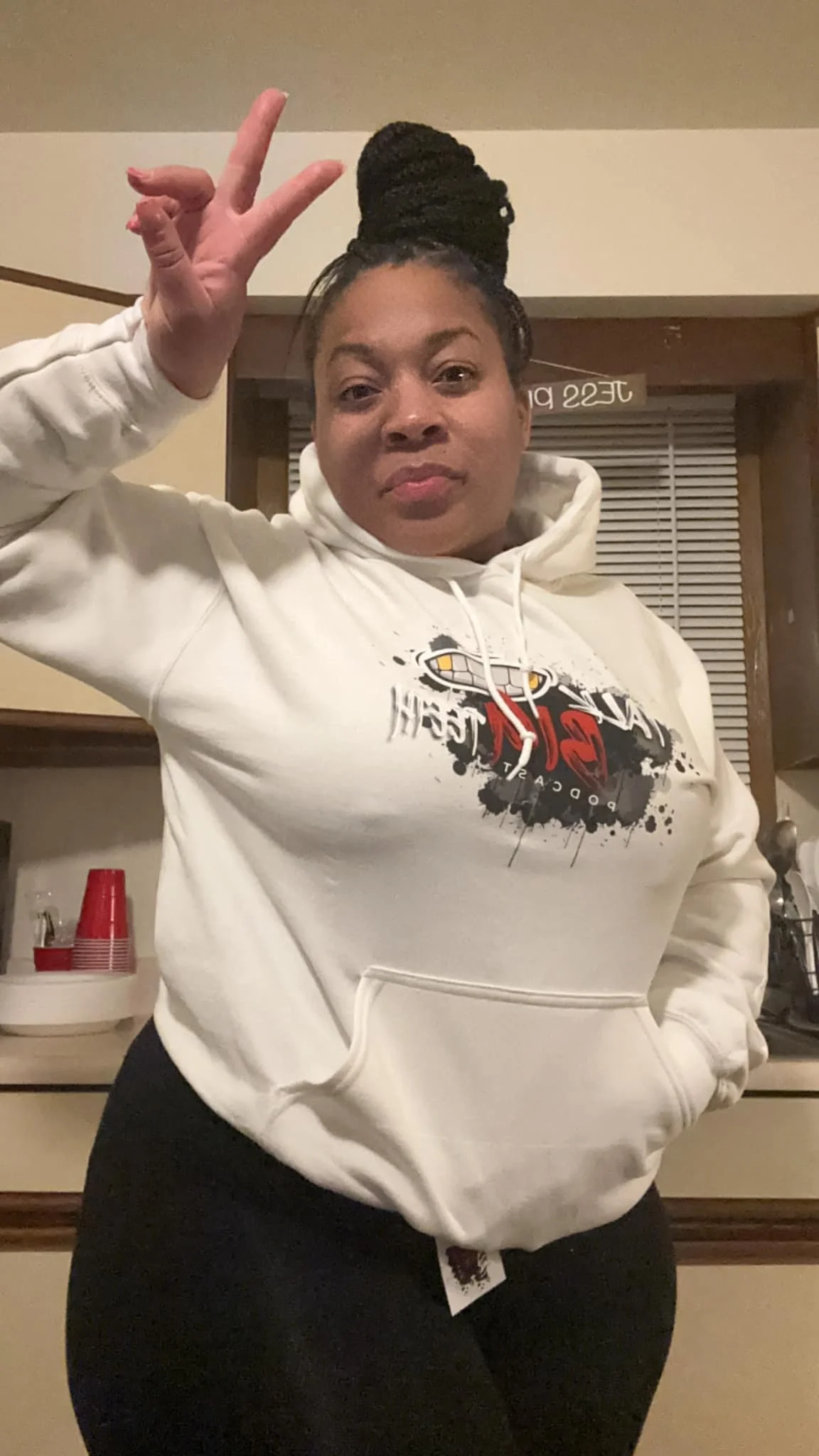 A woman with braided hair wearing a white hoodie and black pants, making a peace sign and smiling at the camera indoors.