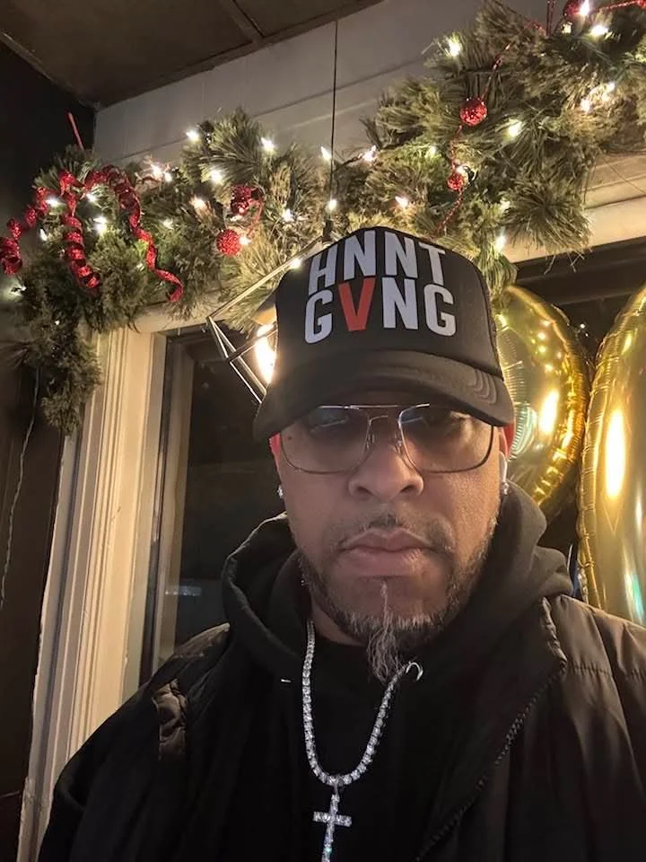 A man wearing a black cap with the words 'HNT G VNG' edited to say 'HATE GIVING', glasses, a black hoodie, and a silver cross necklace. He is inside a room decorated with Christmas garland and lights.