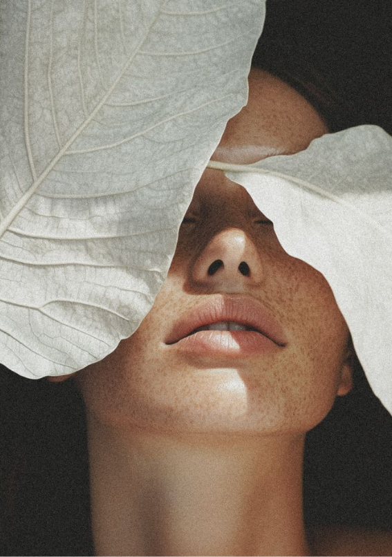 Close-up of a woman with freckled skin and closed eyes, partially covered by large white leaves.