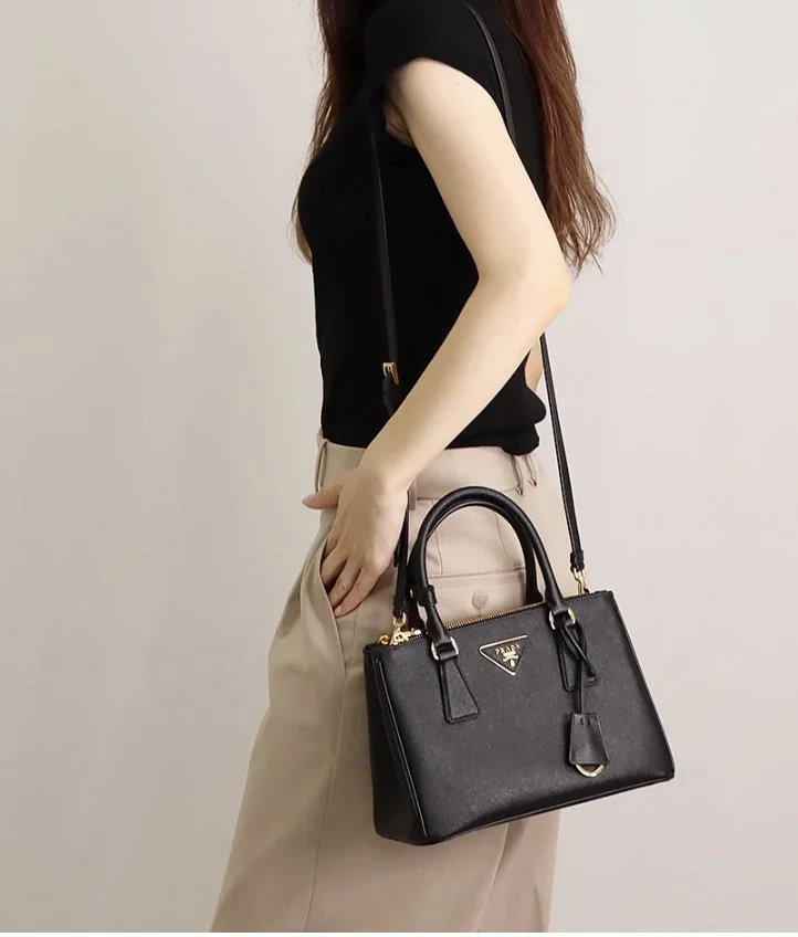 A woman wearing a black sleeveless top and beige pants, carrying a black Prada handbag with gold hardware, standing against a plain background.