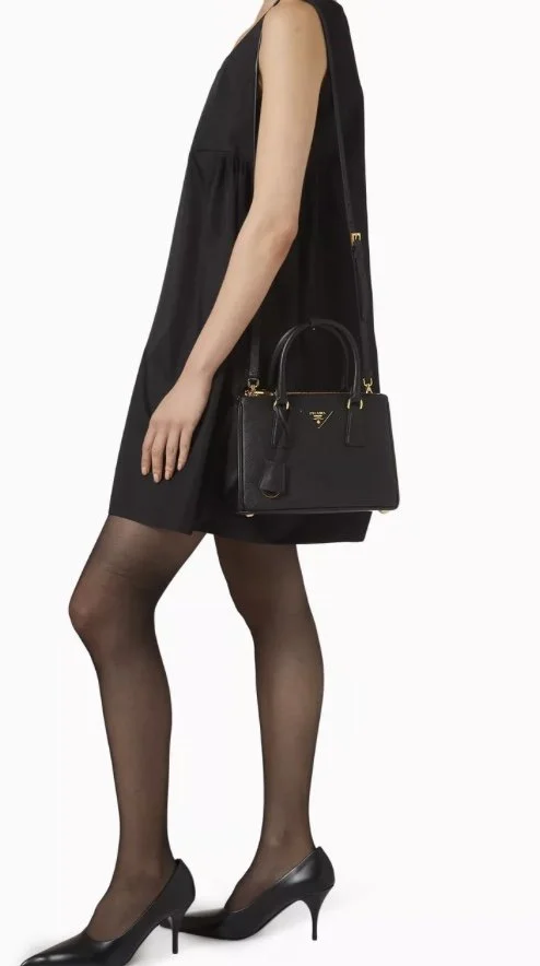 A woman in black dress wearing black stockings and high heels, holding a black Prada handbag.