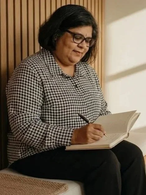 Woman with glasses, black hair, wearing a checkered shirt, sitting and writing in a notebook.