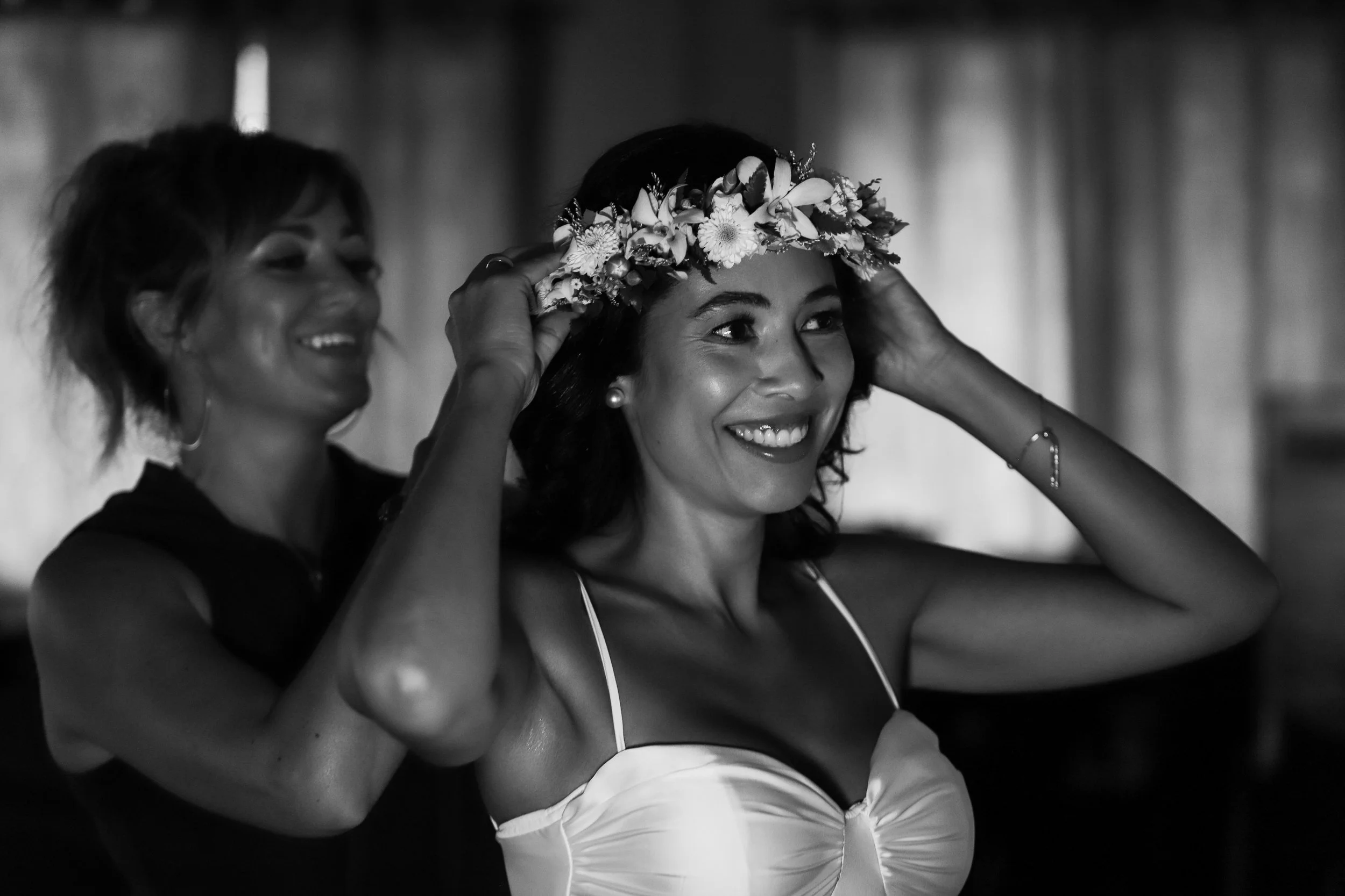 A woman wearing a flower crown is smiling as another woman helps her adjust it, in a black and white photo.