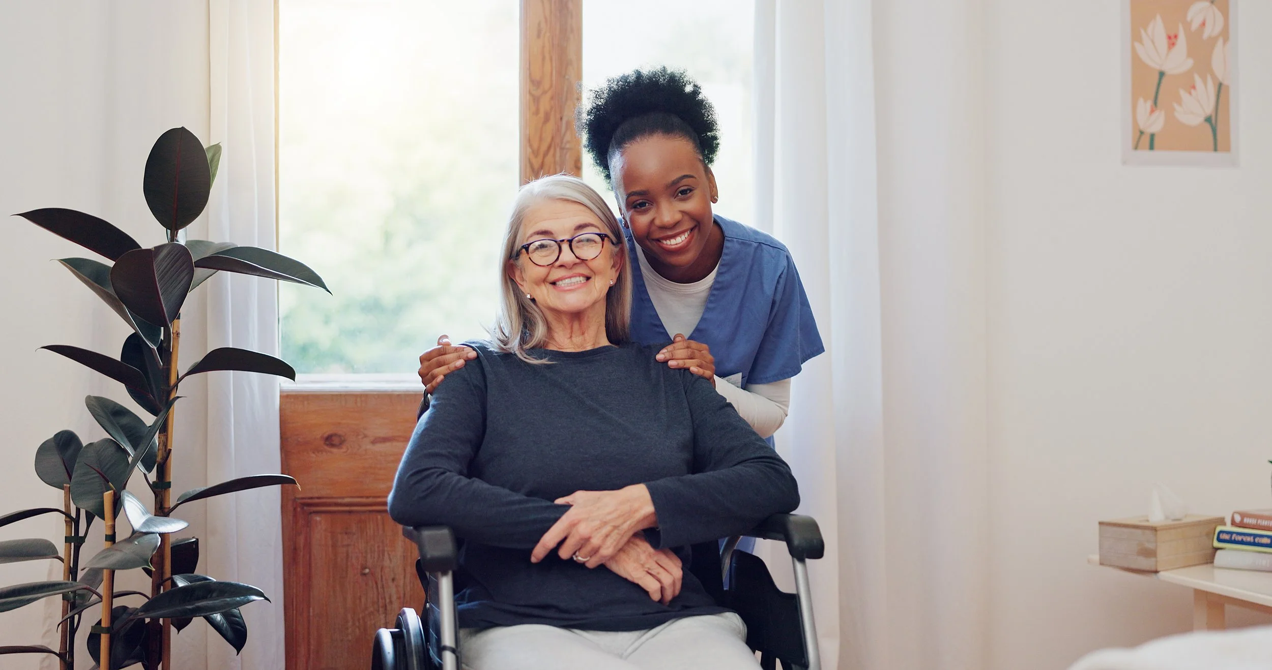 A smiling elderly woman in a wheelchair with gray hair and glasses, accompanied by a young female healthcare worker with dark skin and curly hair, both posing happily indoors near a window with greenery outside.