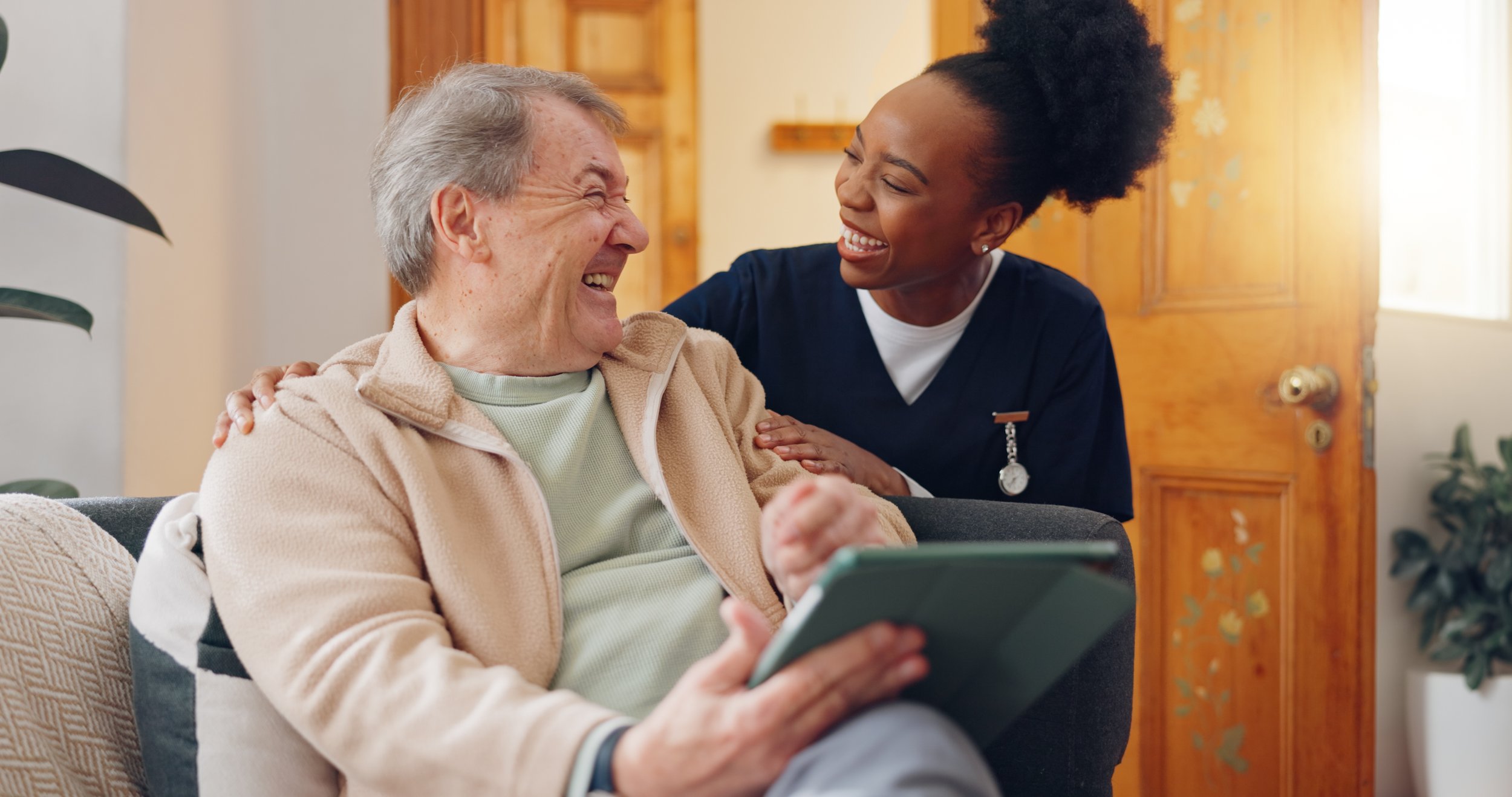 A happy elderly man and a caregiver sharing a laugh inside a warmly lit room, with the man holding a digital tablet.