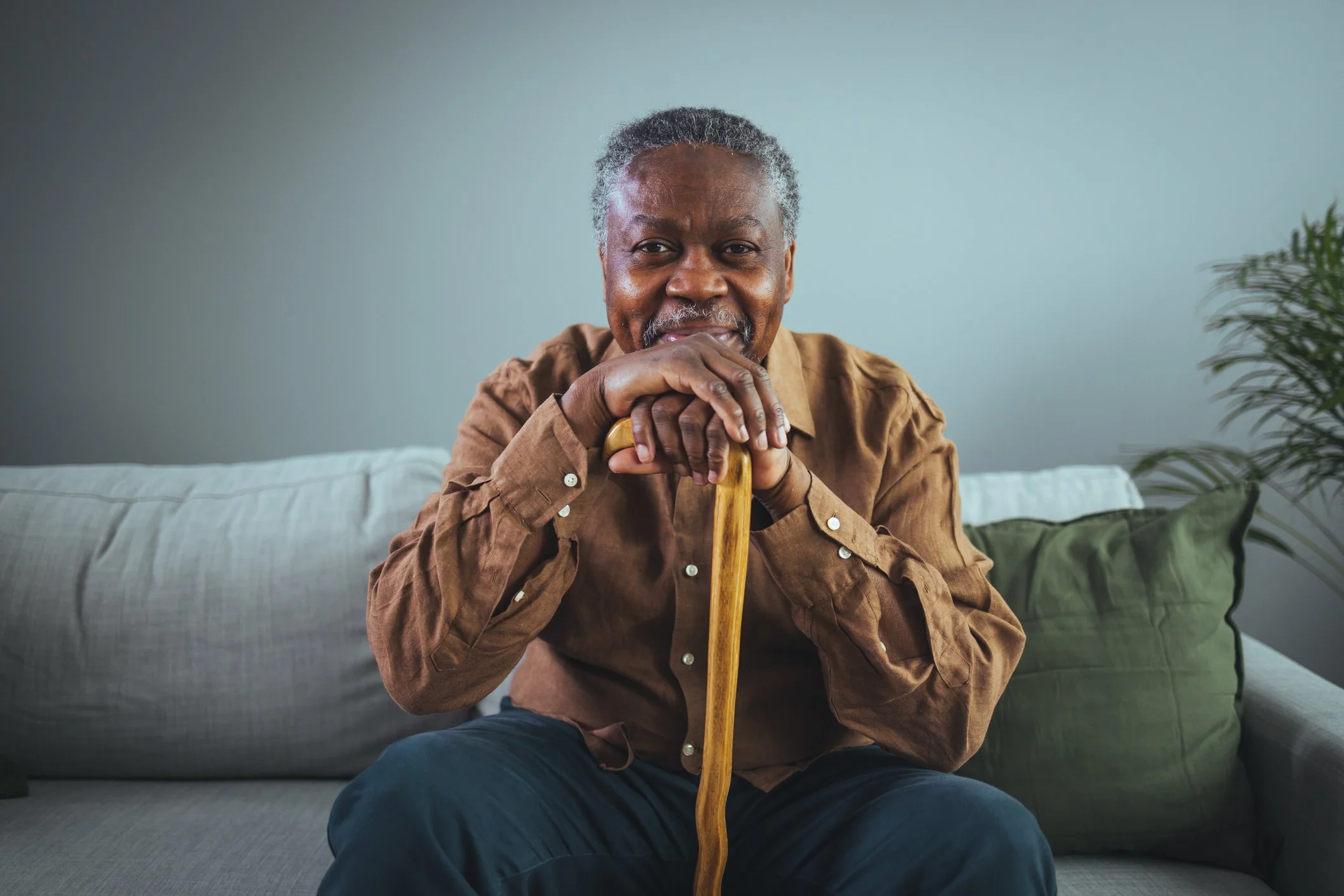 An elderly man with gray hair and a beard sitting on a beige sofa, smiling, with his hands resting on a wooden cane in front of him. The background has a light blue wall and a green potted plant.