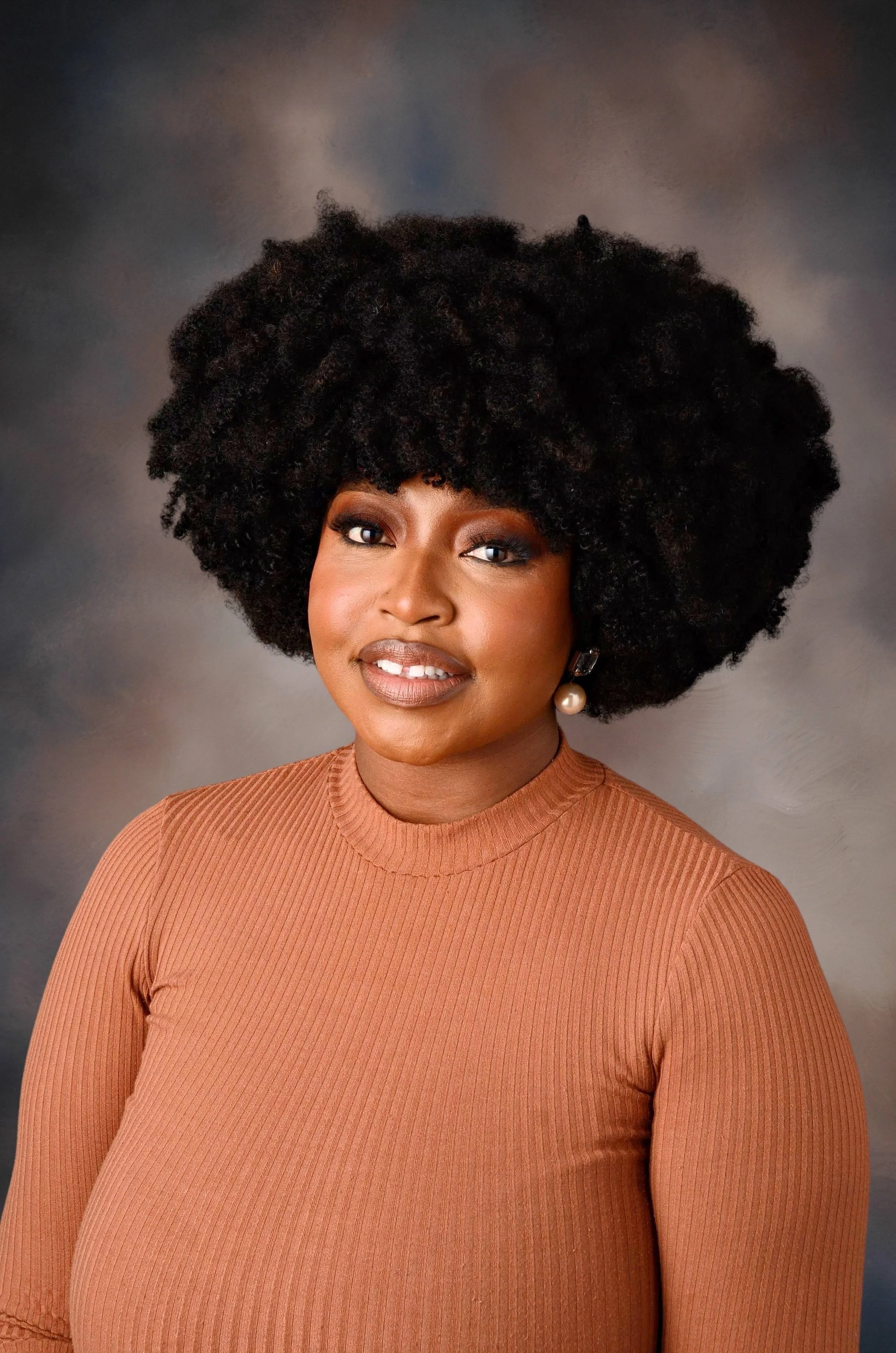 A woman with dark skin and a large, curly black hairstyle. She is wearing a rust-colored, ribbed long-sleeve top and pearl earrings, standing against a neutral, blurred background.
