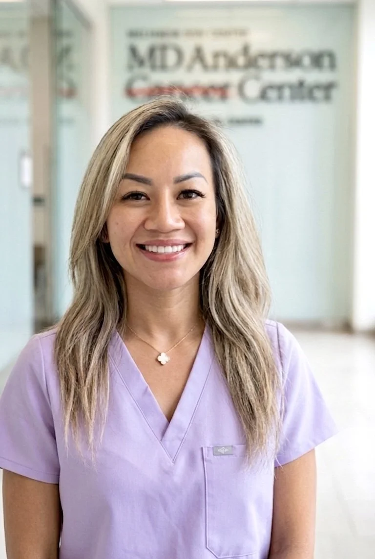 Smiling woman with shoulder-length wavy blonde hair wearing a light purple medical scrub top and a necklace with a four-leaf clover pendant, standing in front of a glass wall with a blurred sign that reads 'MD Anderson Cancer Center'.