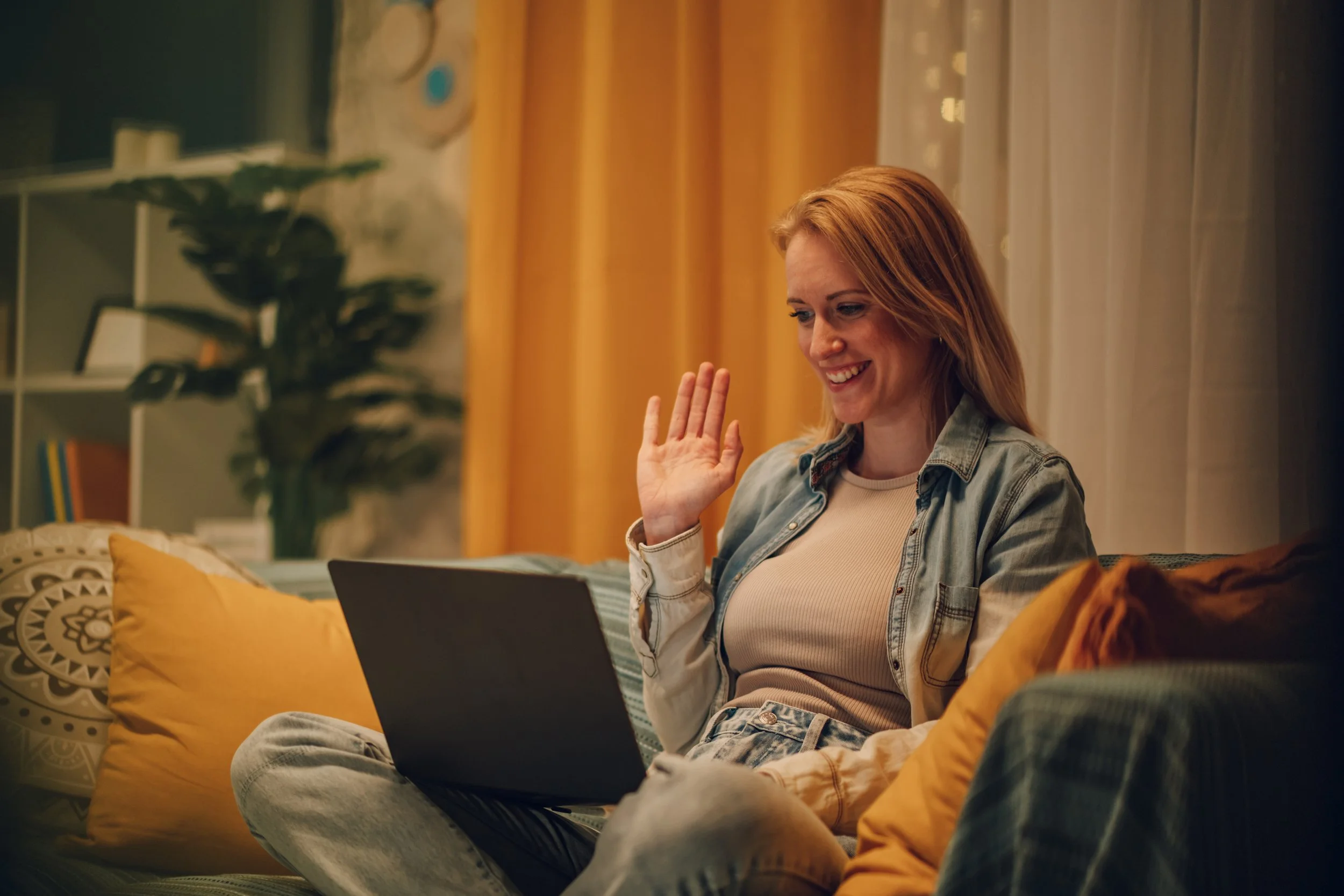 A smiling woman sitting on an inviting living room couch during telehealth therapy. She is waving at her laptop which is placed on her lap.