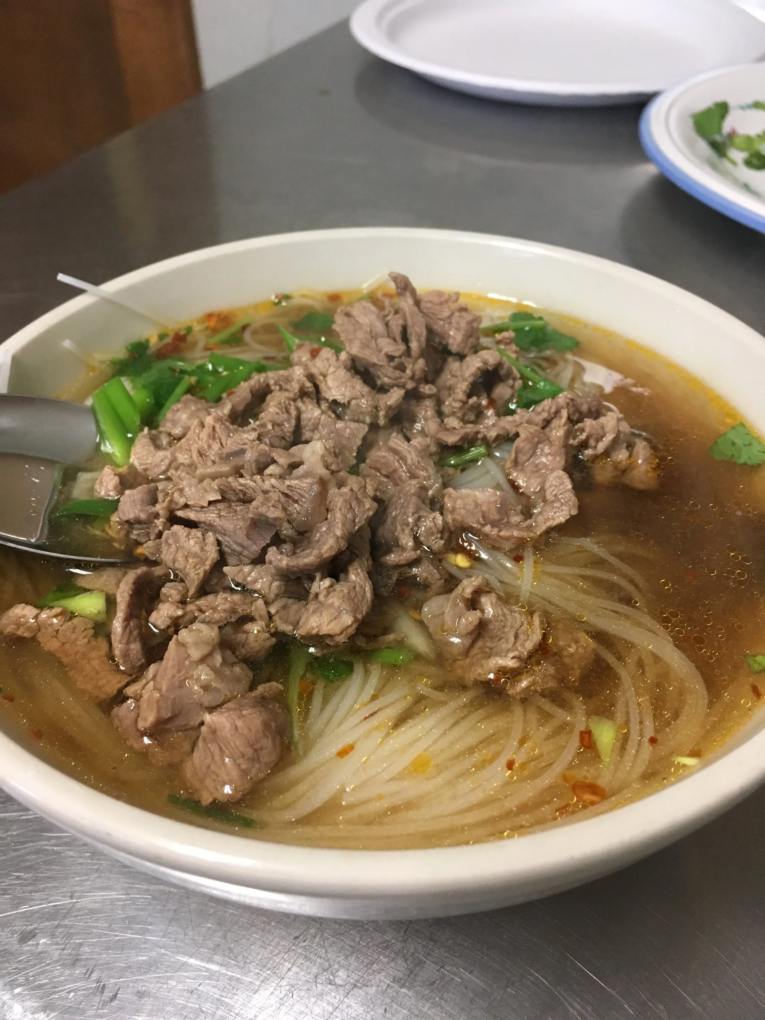 Bowl of beef noodle soup with rice noodles, sliced beef, cilantro, green onions, and broth on a metal table.