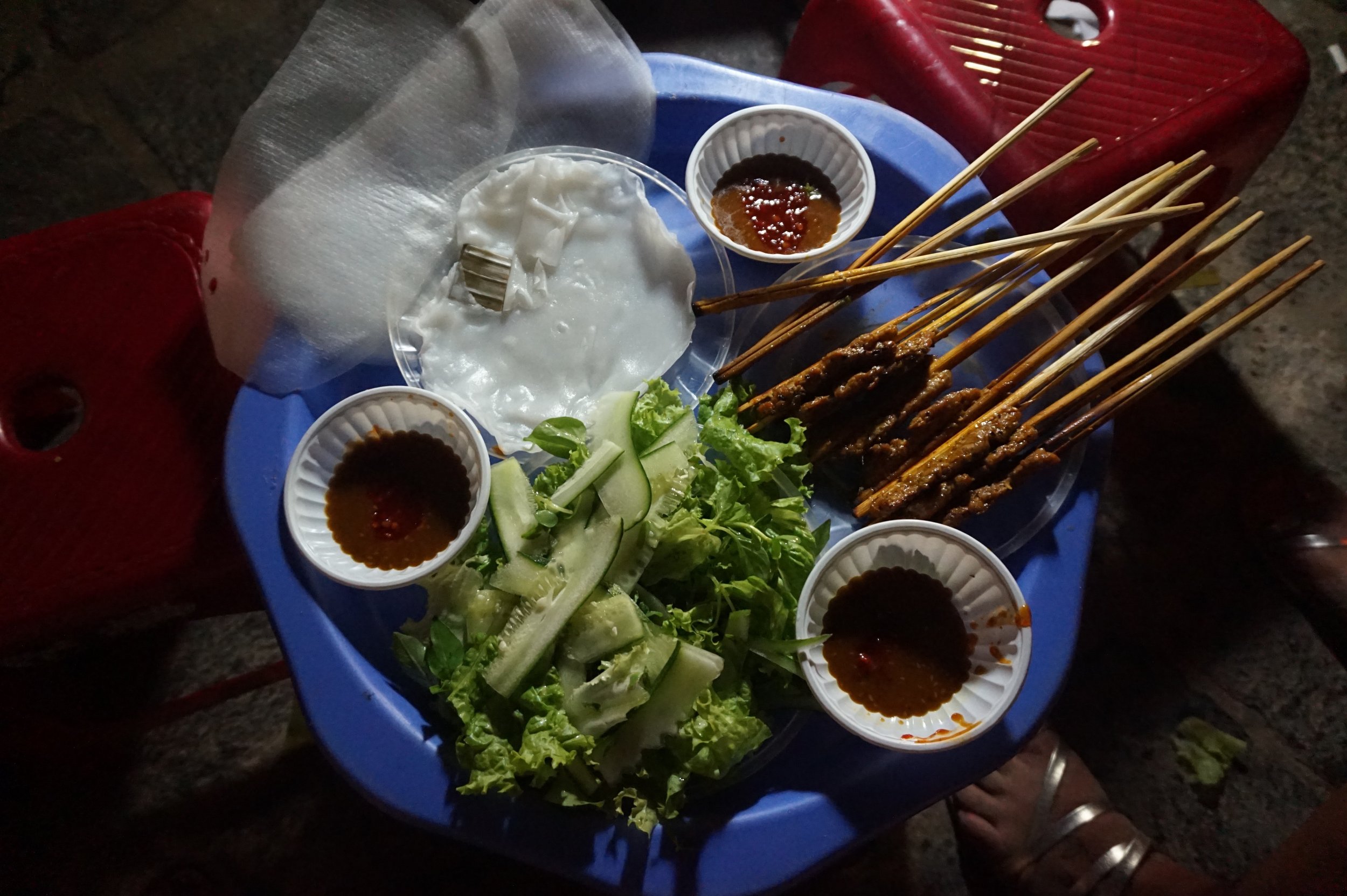 Street food platter with grilled meat skewers, lettuce, cucumber, rice paper, and sauces.