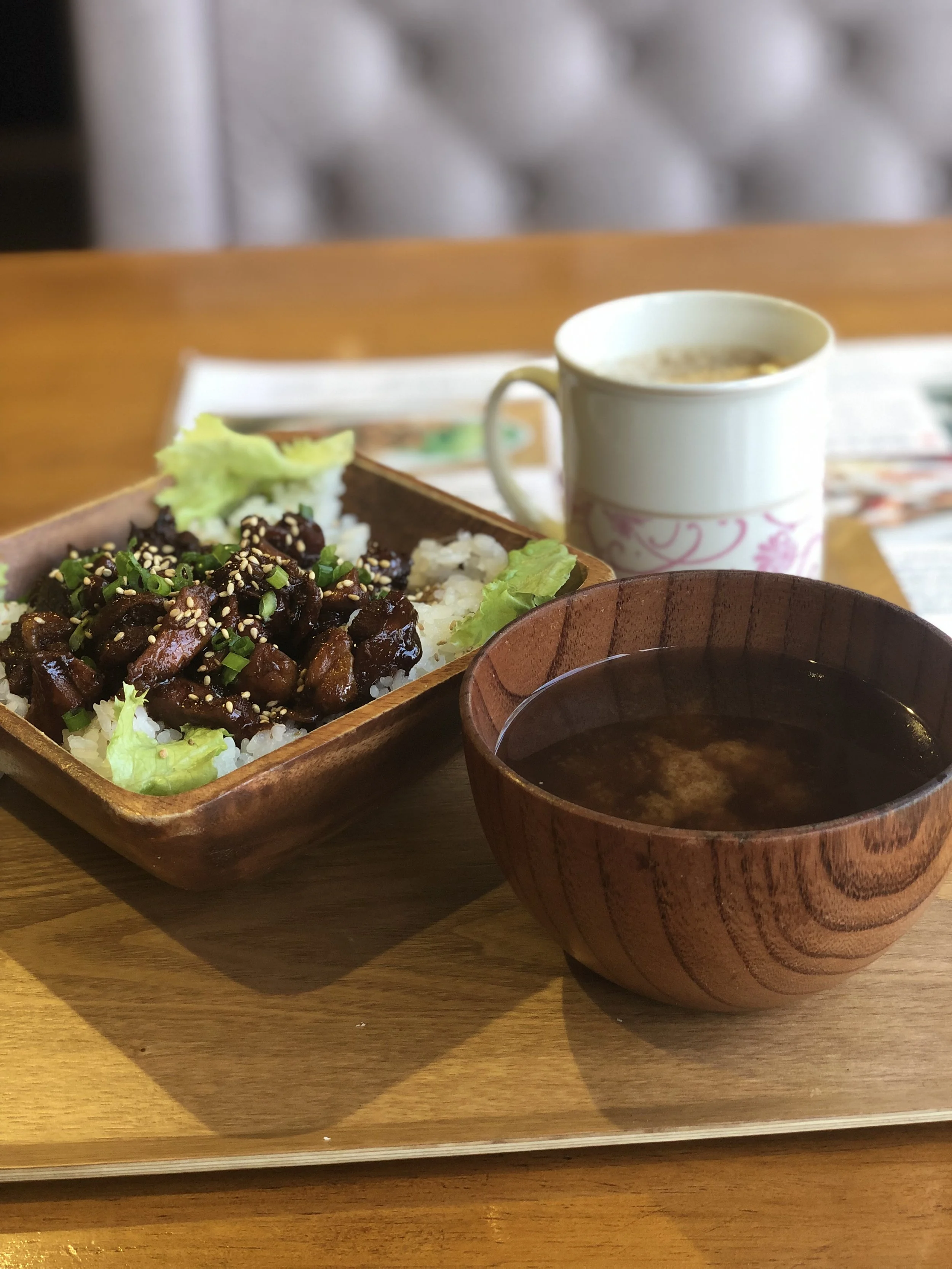 A wooden tray holding a bowl of miso soup, a rice bowl with meat topped with sesame seeds, and a mug of coffee on a wooden table.