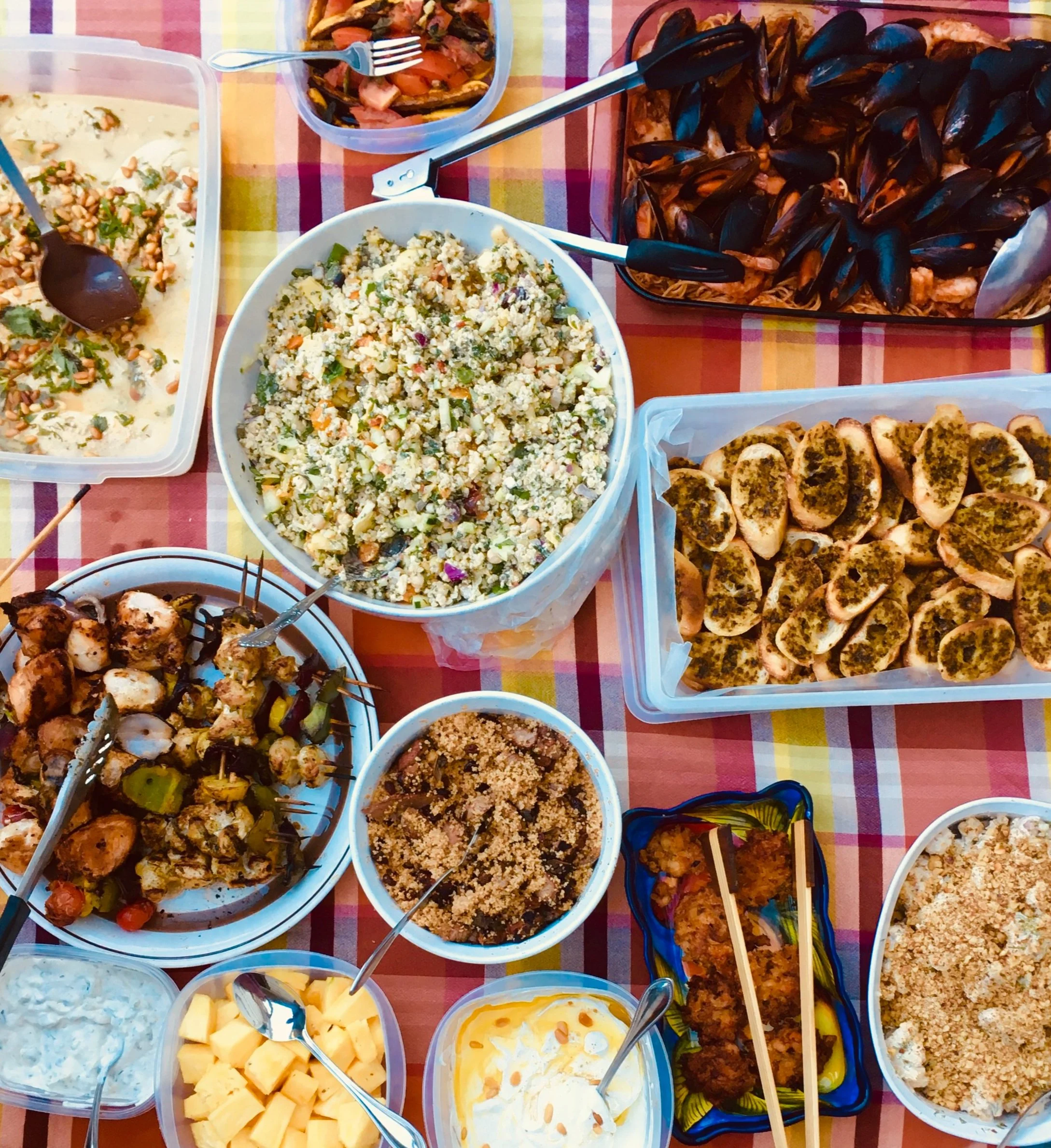 A variety of dishes on a colorful tablecloth, including mixed salads, skewers, mussels, seasoned bread slices, and assorted dips.