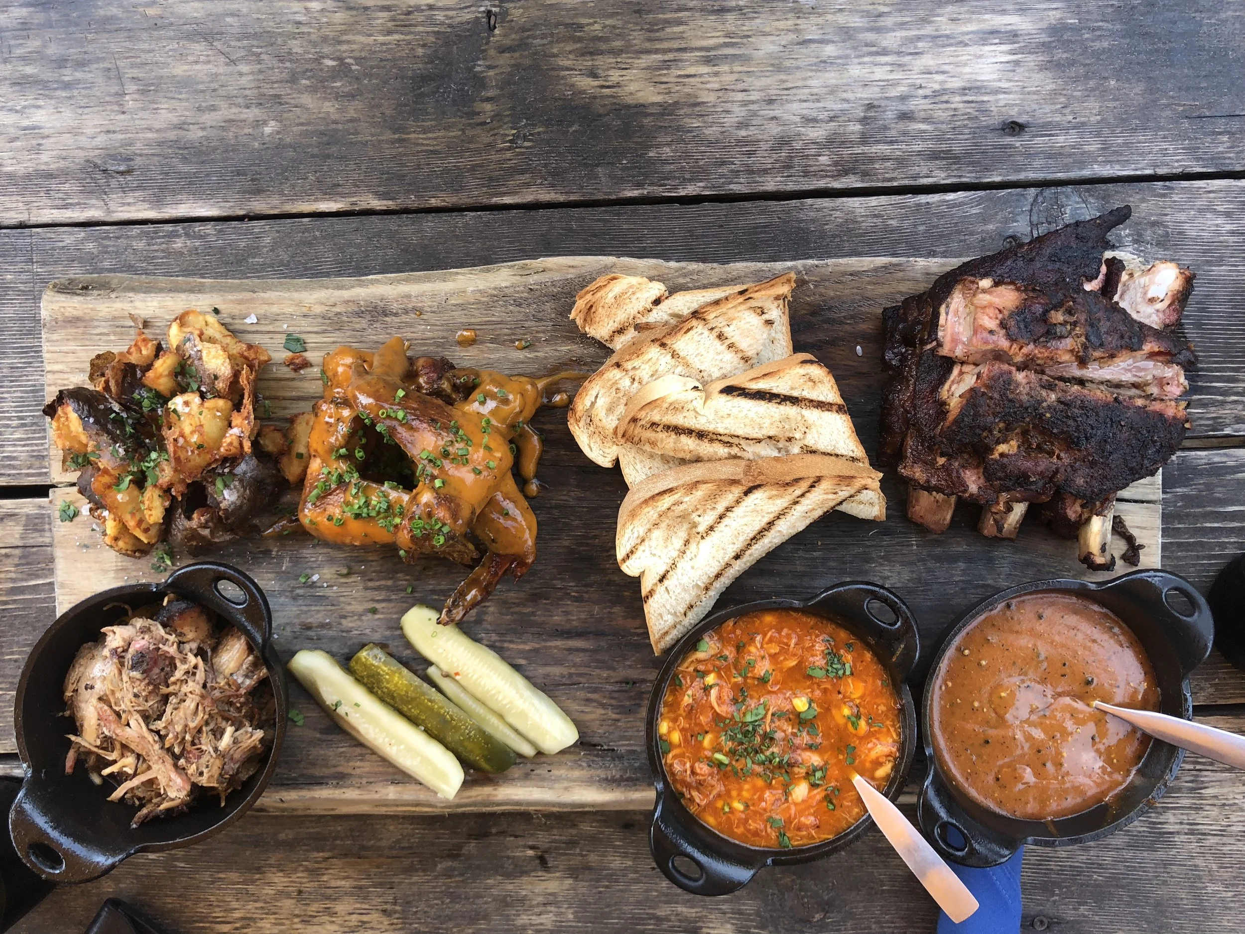 Barbecue platter with ribs, grilled bread, saucy chicken wings, roast potatoes, pulled pork in a skillet, pickles, baked beans, and sauce in small pots on a wooden table.