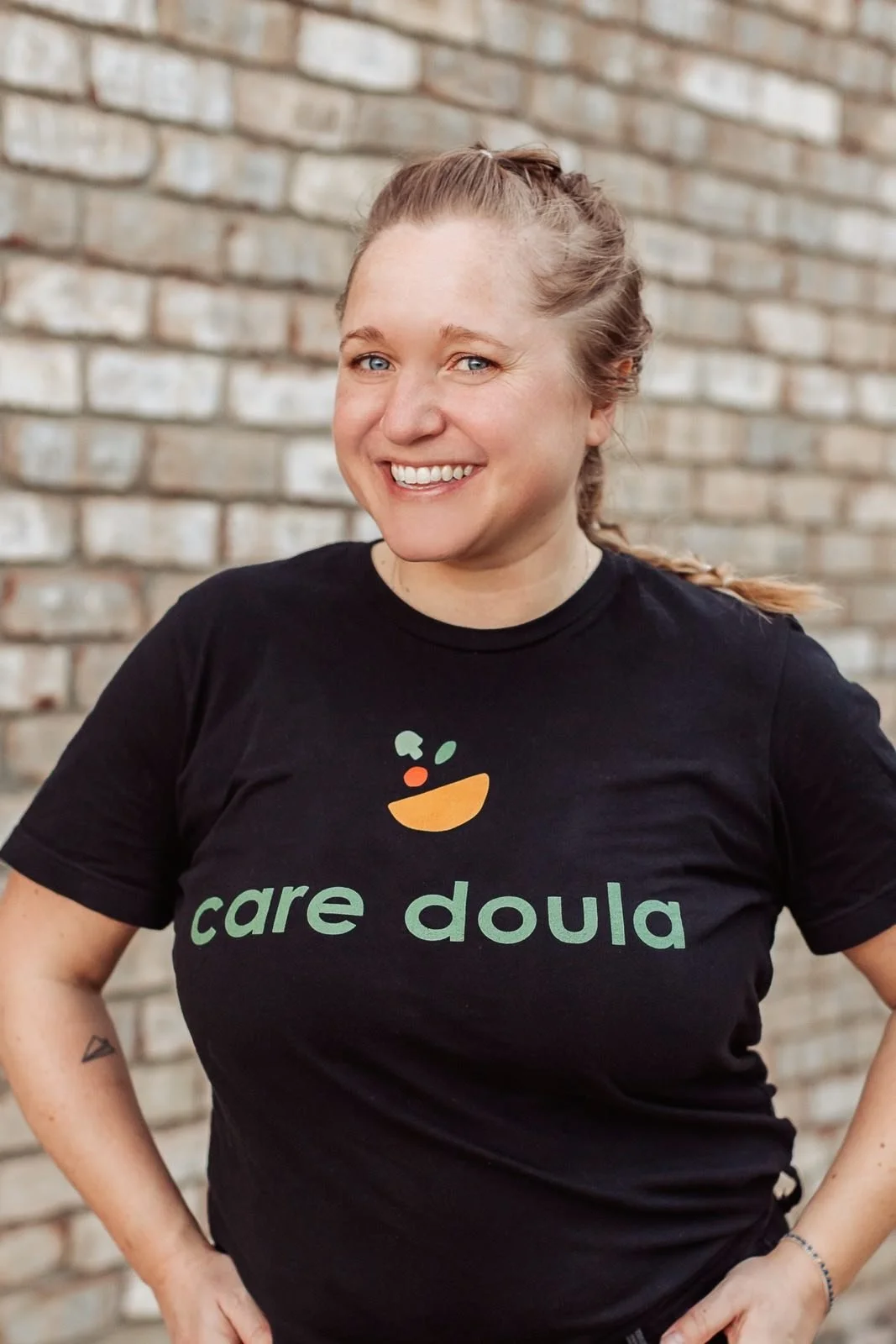A smiling woman with blonde hair tied back, wearing a black t-shirt with the words 'care doula' and a colorful bowl logo, standing in front of a brick wall.