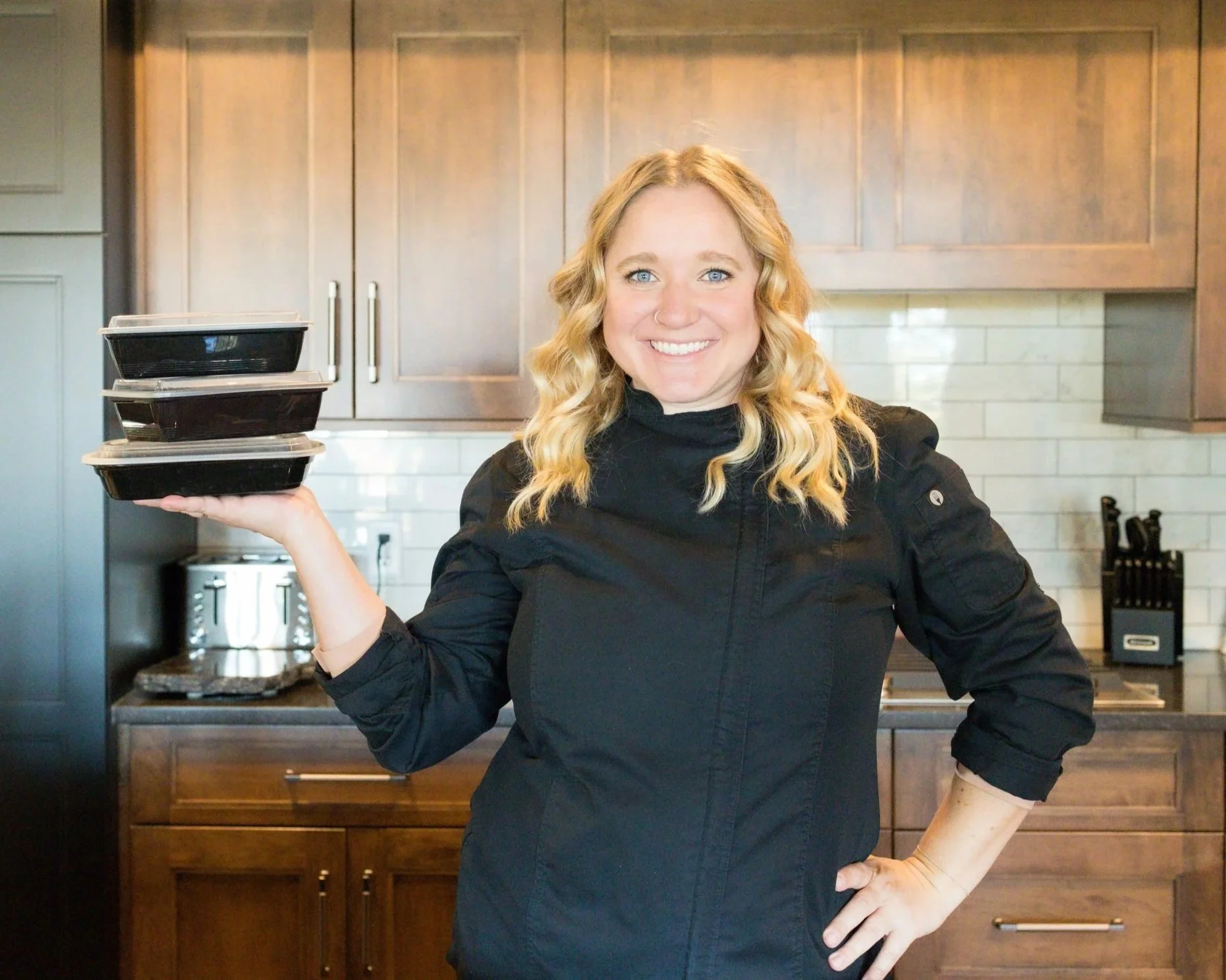 A woman with blonde curly hair in a black chef's coat smiling while holding three takeout containers in a kitchen with wooden cabinets and white tile backsplash.