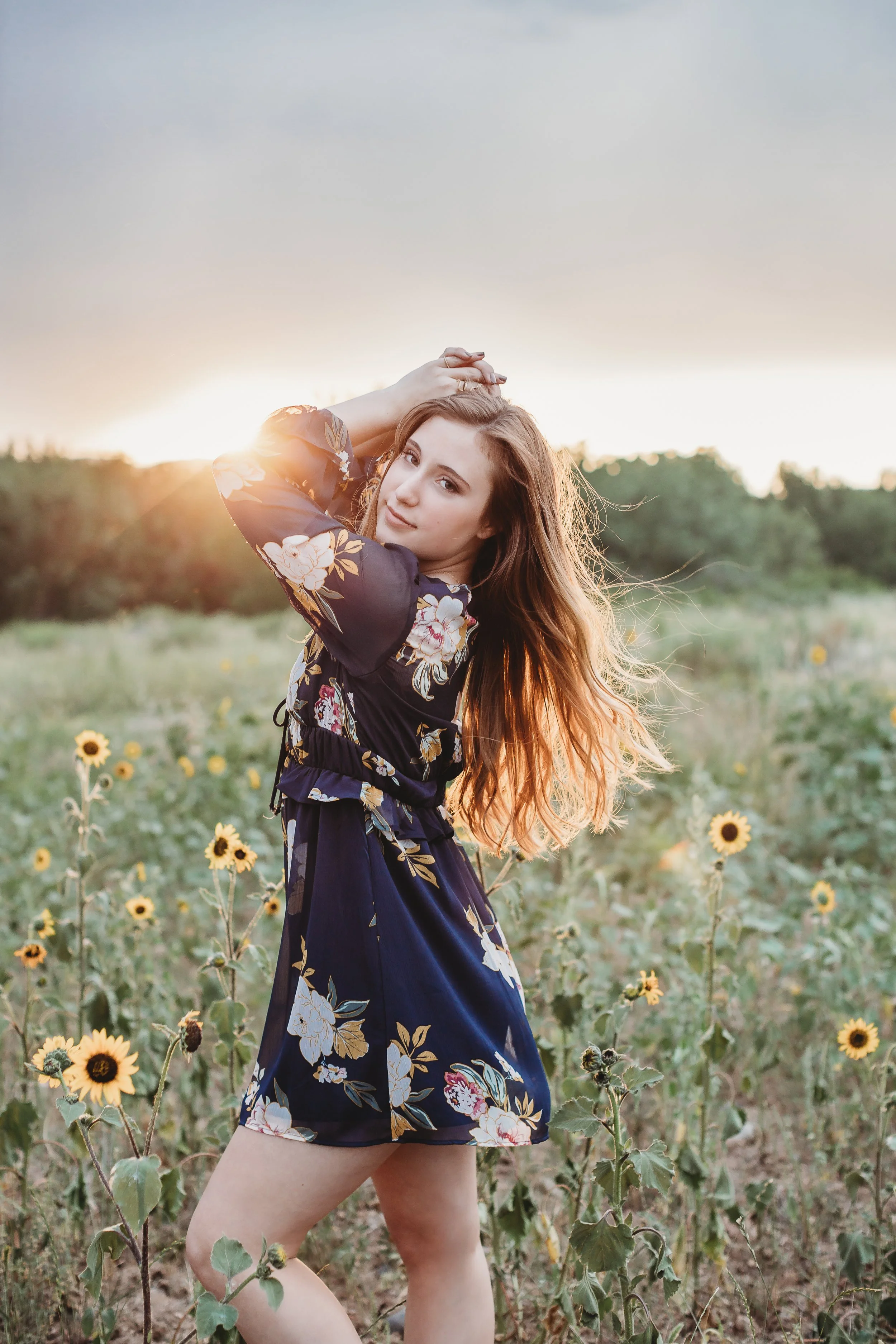 A young woman with long, wavy hair standing in a sunflower field at sunset, wearing a dark floral dress, with her hands raised to her head, looking at the camera.