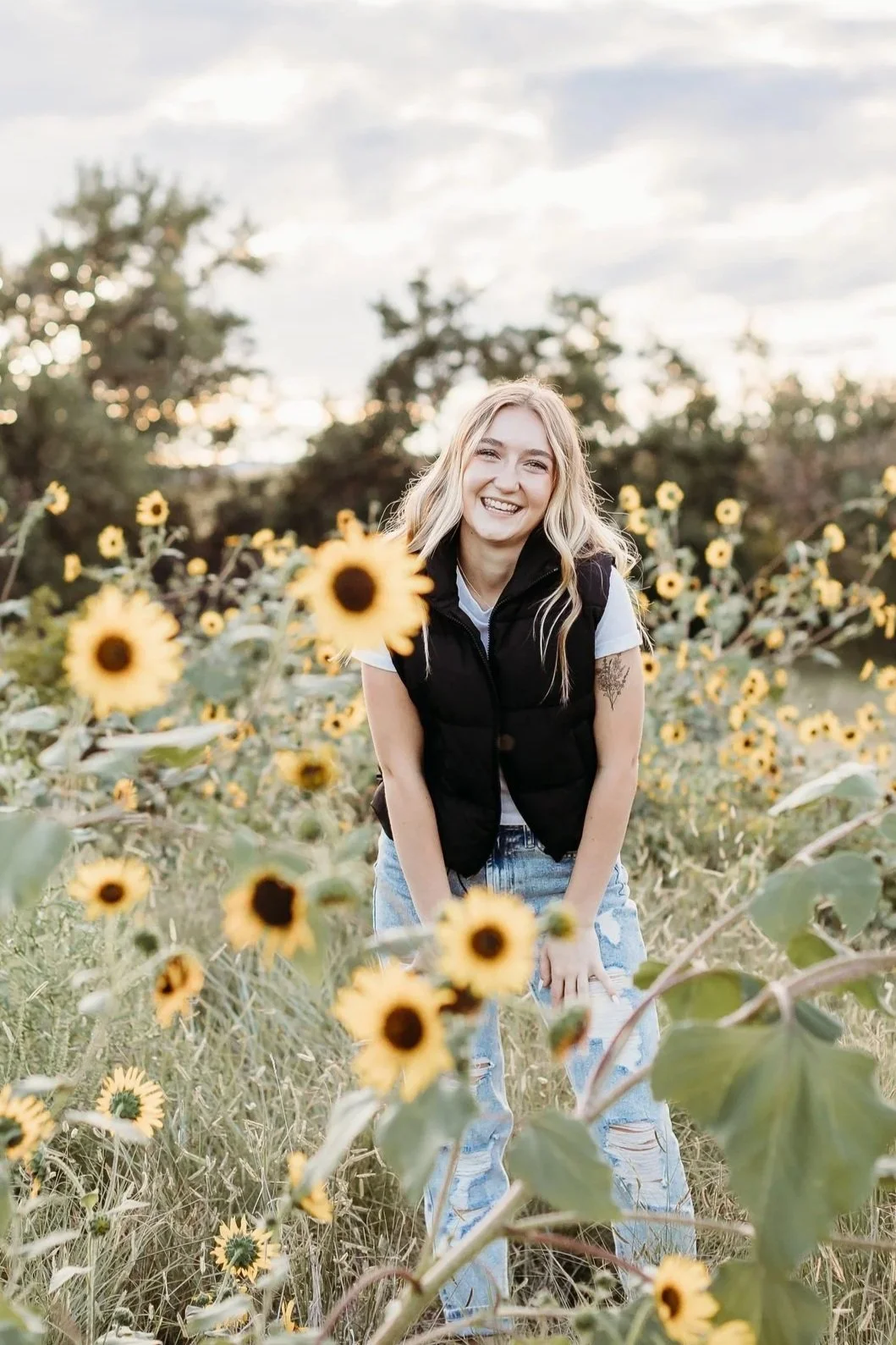 A young woman with blonde hair, smiling and leaning forward in a field of sunflowers during sunset.